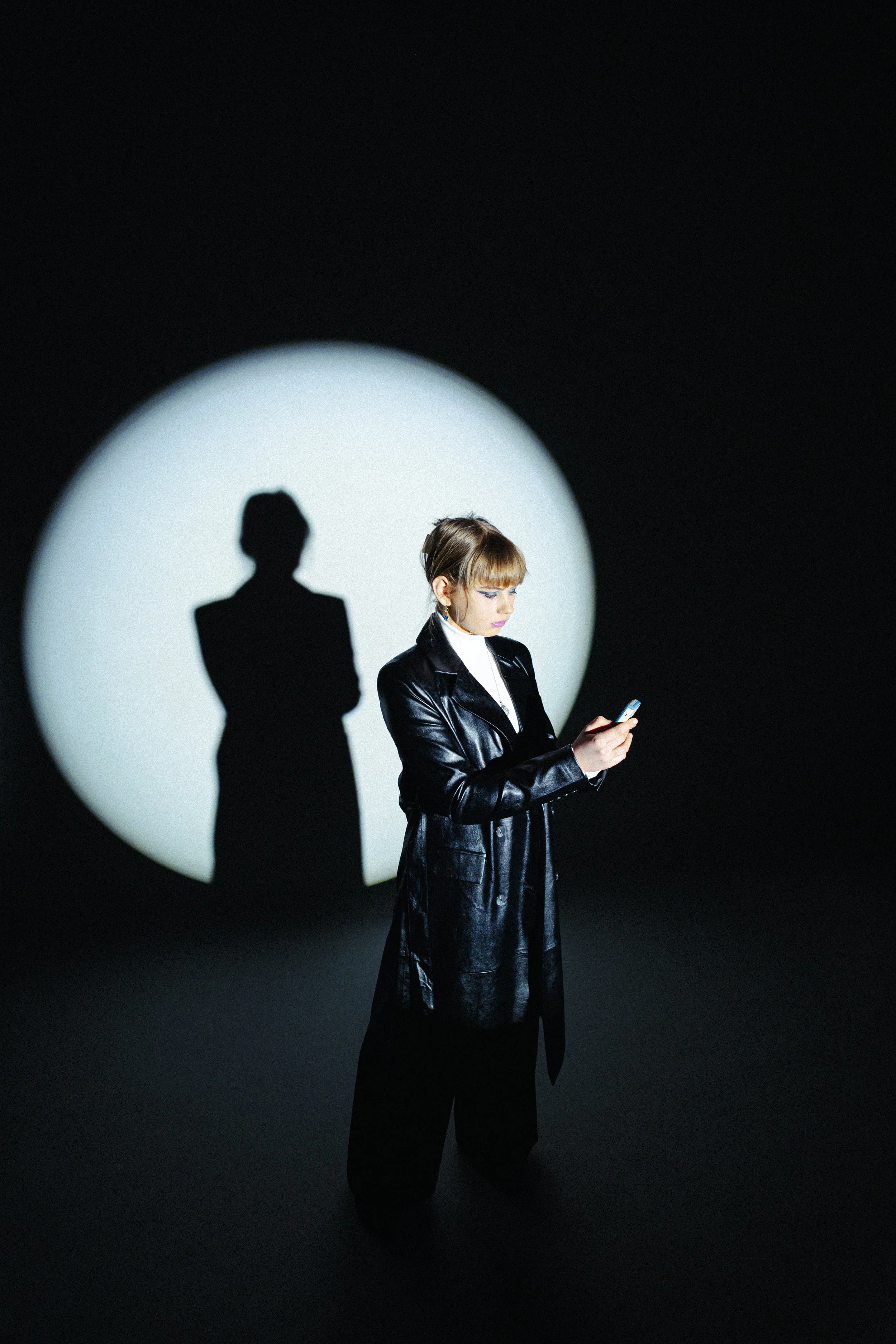 A woman with short hair, blue eye makeup, and large earrings is standing against a black background with a white spotlight. Her shadow is cast on the wall behind her, and she is looking at her phone.
