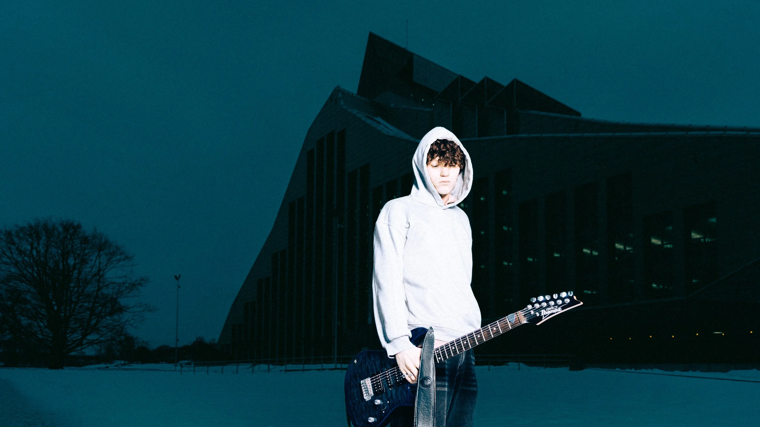 A young person with curly hair wearing a light gray hoodie, holding an electric guitar, standing outdoors at dusk with a modern building in the background and trees in the distance.