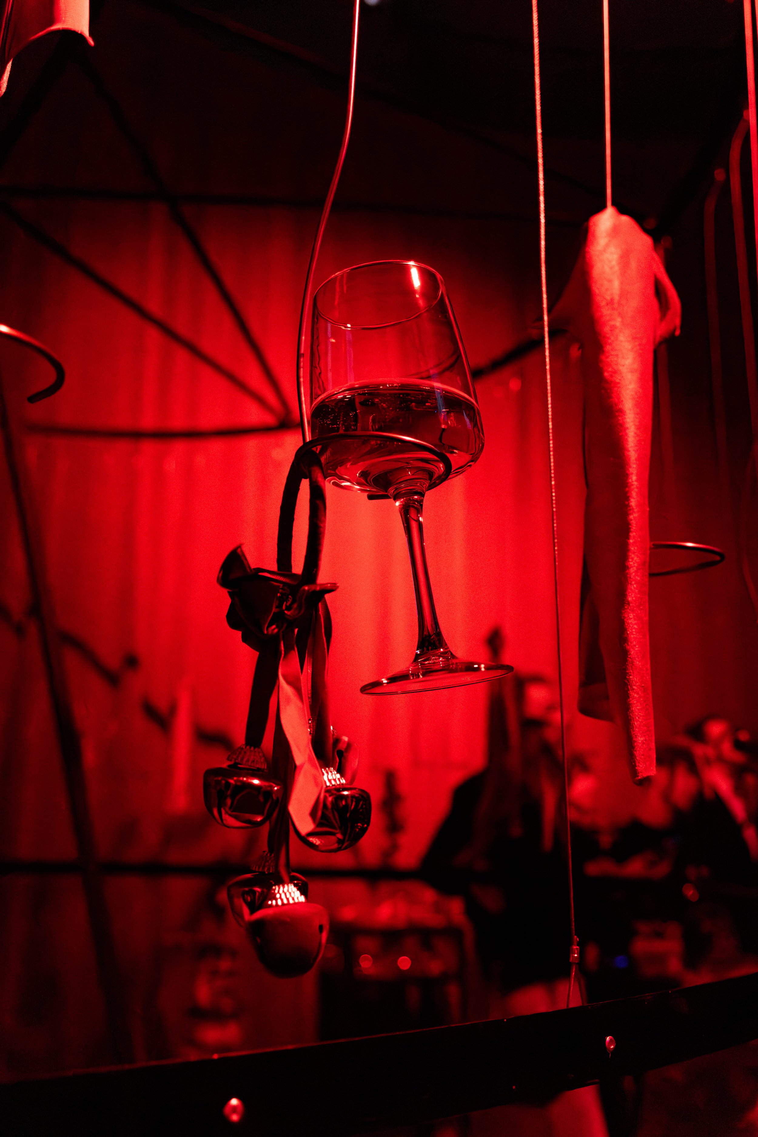 A wine glass filled with red liquid hanging from a thin string against a red-lit background with fabric and metal hooks.