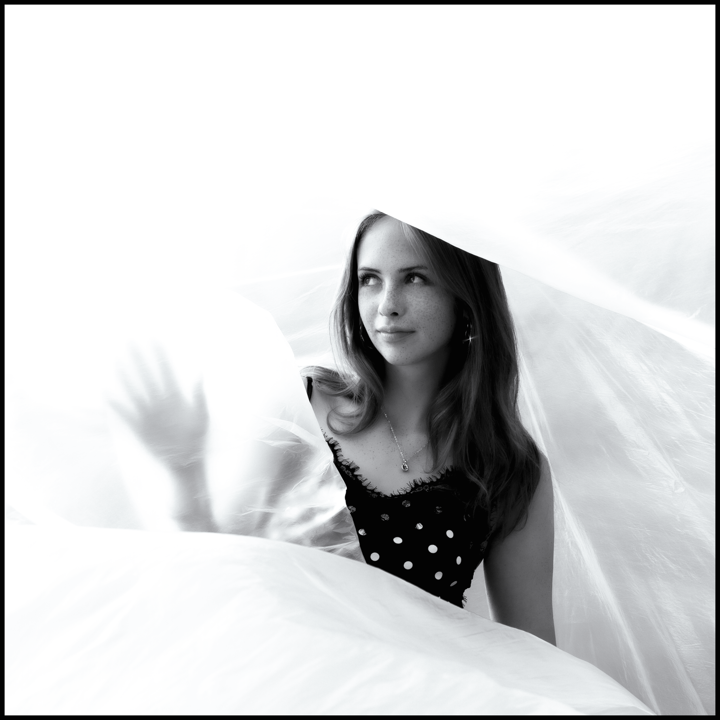 Black and white portrait of a young woman with long hair, wearing a polka dot top and a necklace, looking to the side, partially obscured by an overhang.