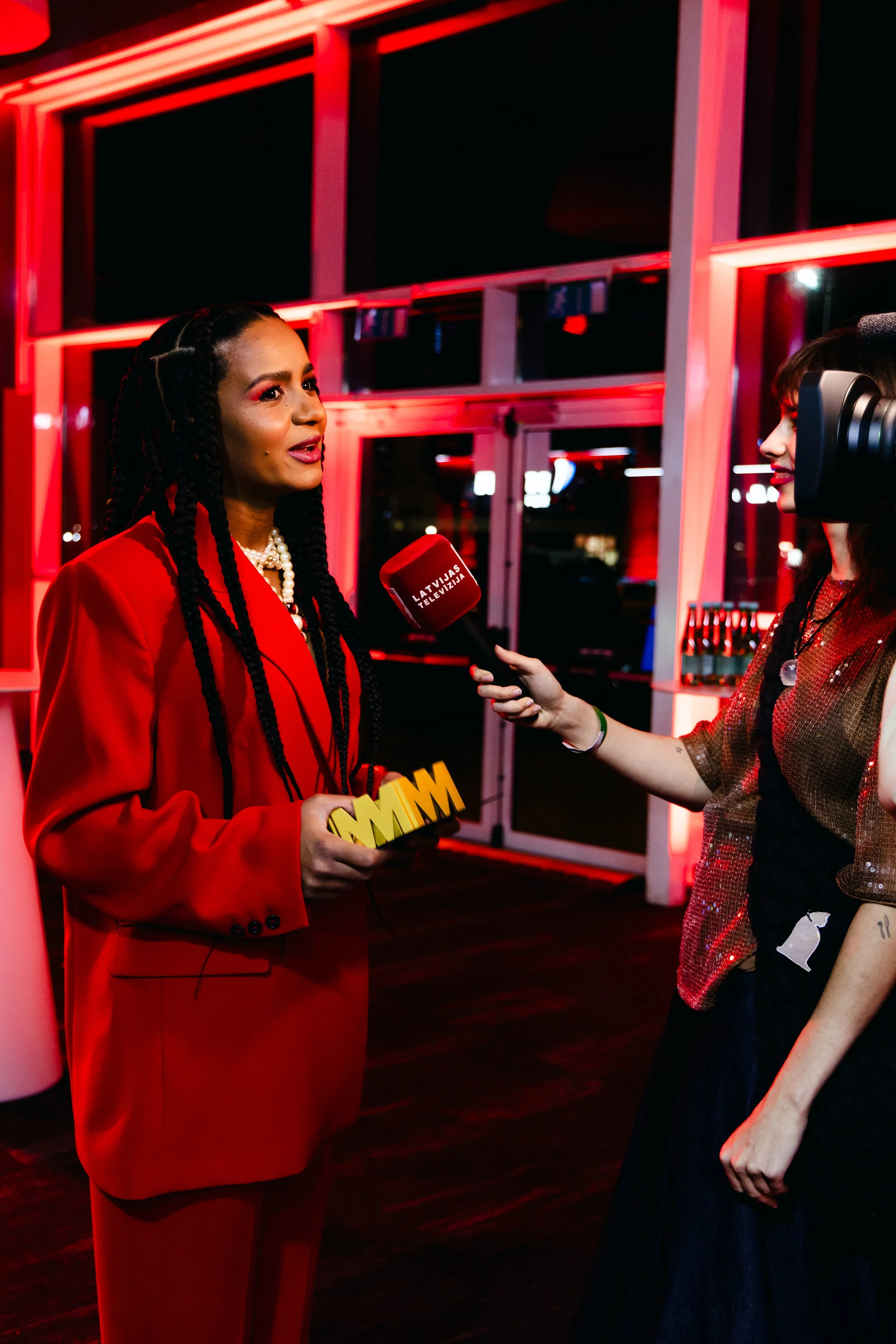 Woman with long braided hair wearing a red suit and pearls being interviewed with a microphone that reads "Latvijas Televizija" by another woman with dark hair in a sparkly top at night inside a building with red neon lighting.