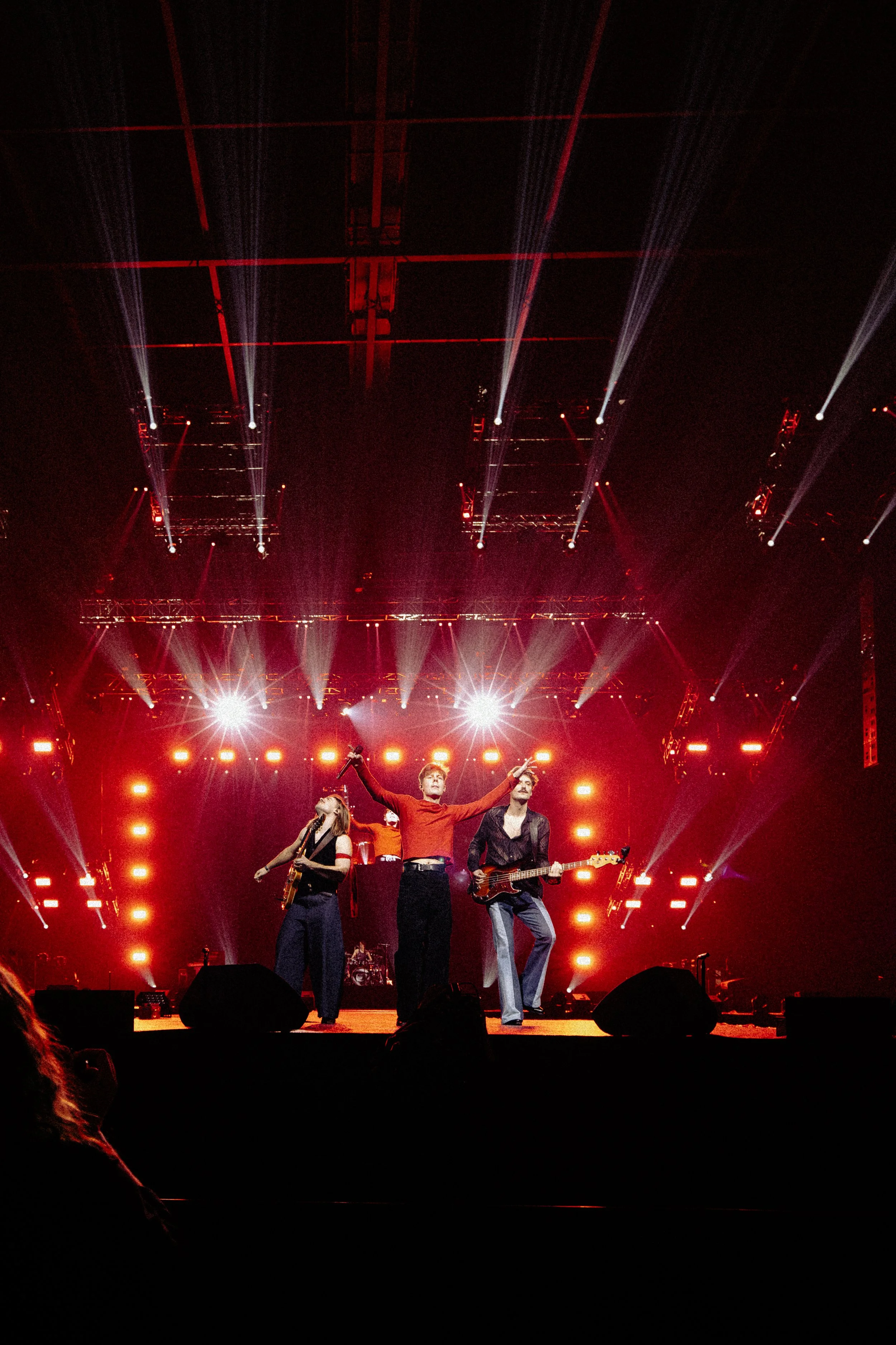 A band performing on stage with three musicians. The stage is lit with red and white lights, and there are beams of light streaking across the ceiling in a concert setting.