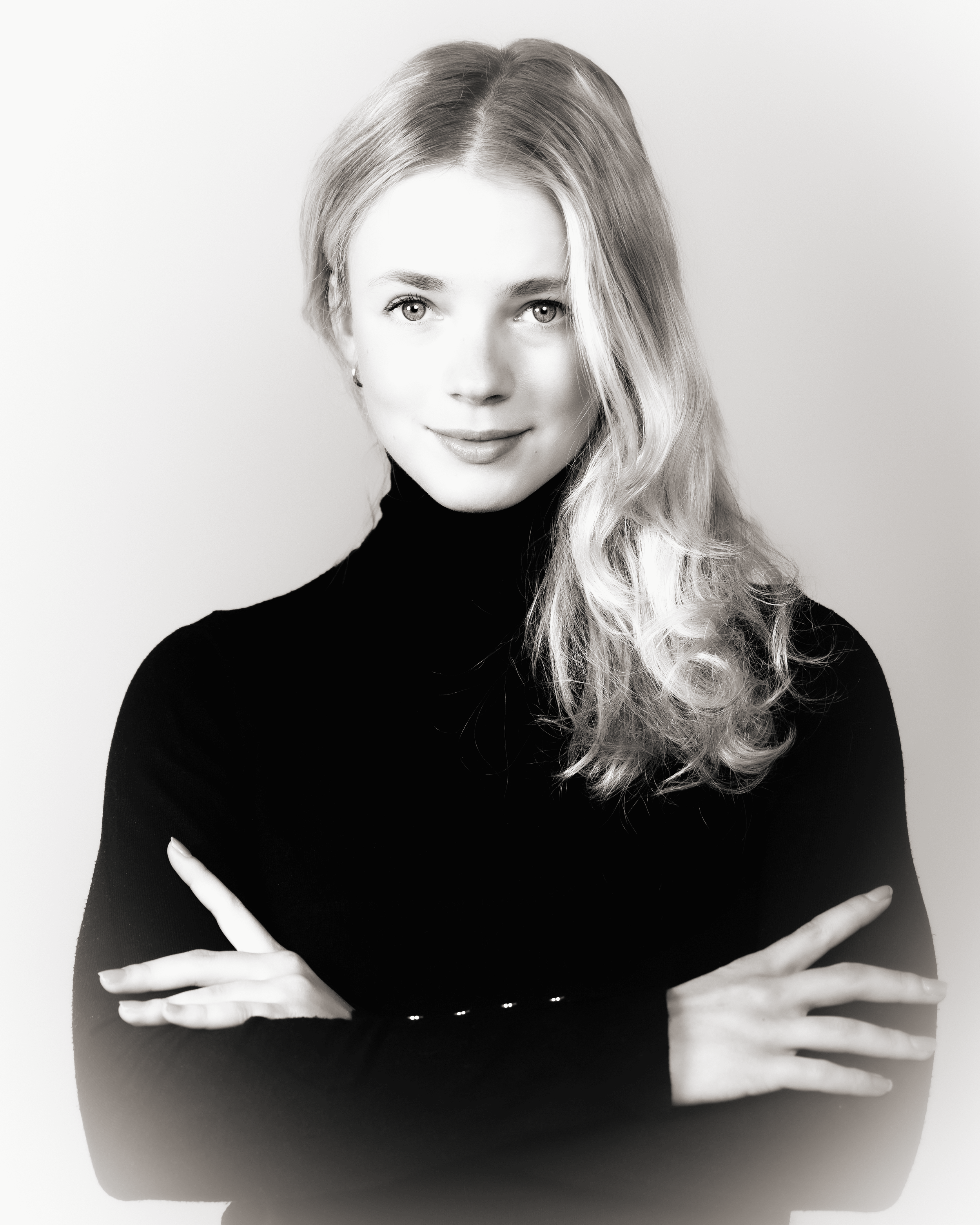 Black and white portrait of a woman with long wavy hair, wearing a dark turtleneck, crossing her arms, looking directly at the camera with a slight smile.