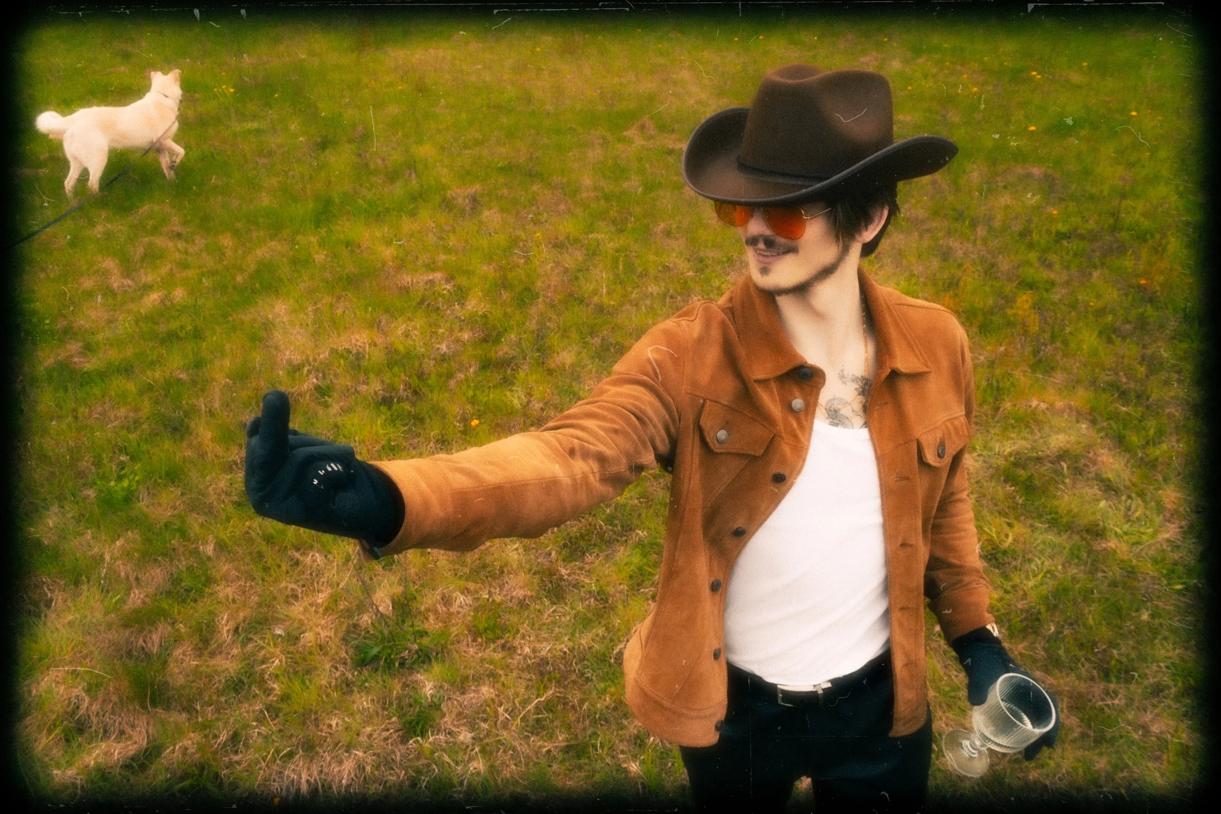 A man wearing a black wide-brimmed hat, orange sunglasses, a brown jacket, and a white shirt is taking a selfie in a grassy field. He is giving a thumbs-up with one hand, and there is a white dog on a leash in the background.