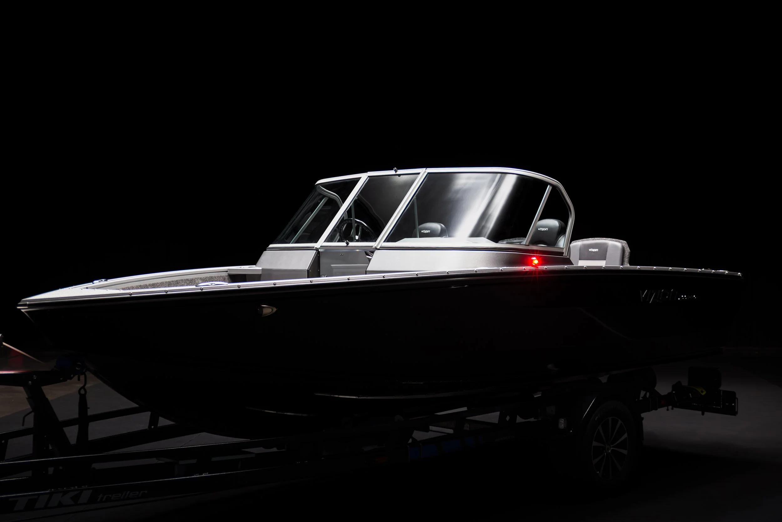 A sleek, modern motorboat with a glass-enclosed cabin, sitting on a trailer against a dark background.