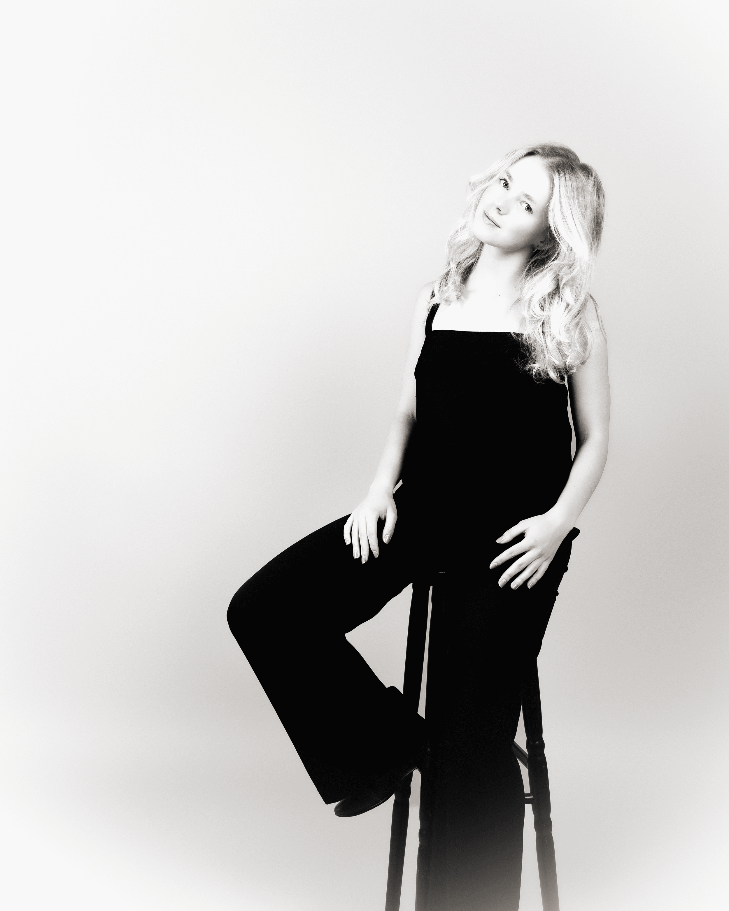 Black and white photograph of a woman with wavy, shoulder-length hair, sitting on a wooden chair against a plain background. She is wearing a sleeveless top and dark pants, with her left leg crossed over her right, and her hands resting on her lap. S