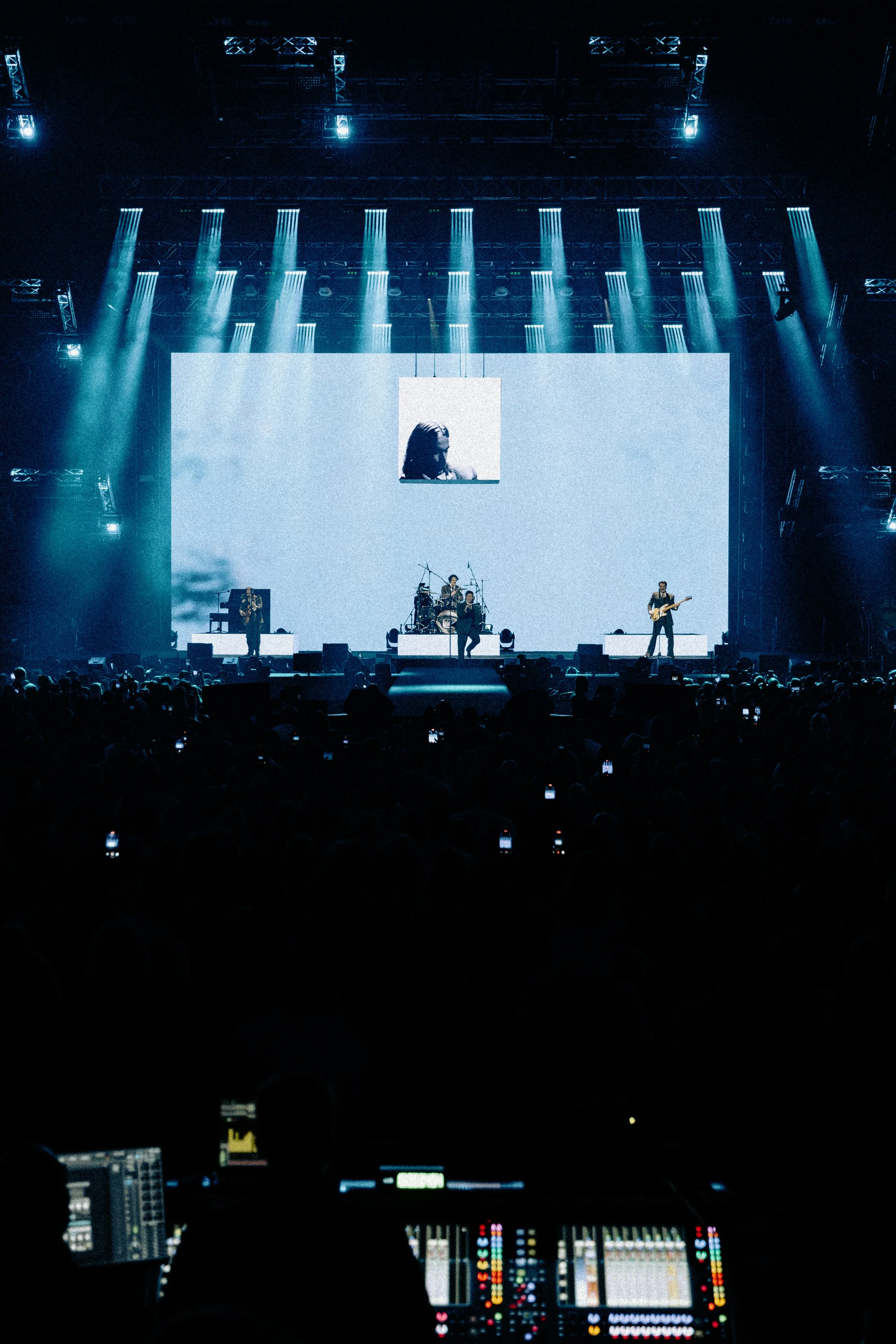 A live concert with a band performing on stage, bright lights, a large LED screen in the background, and an audience in the foreground. The concert is accompanied by a sound engineer or DJ at a mixing console.