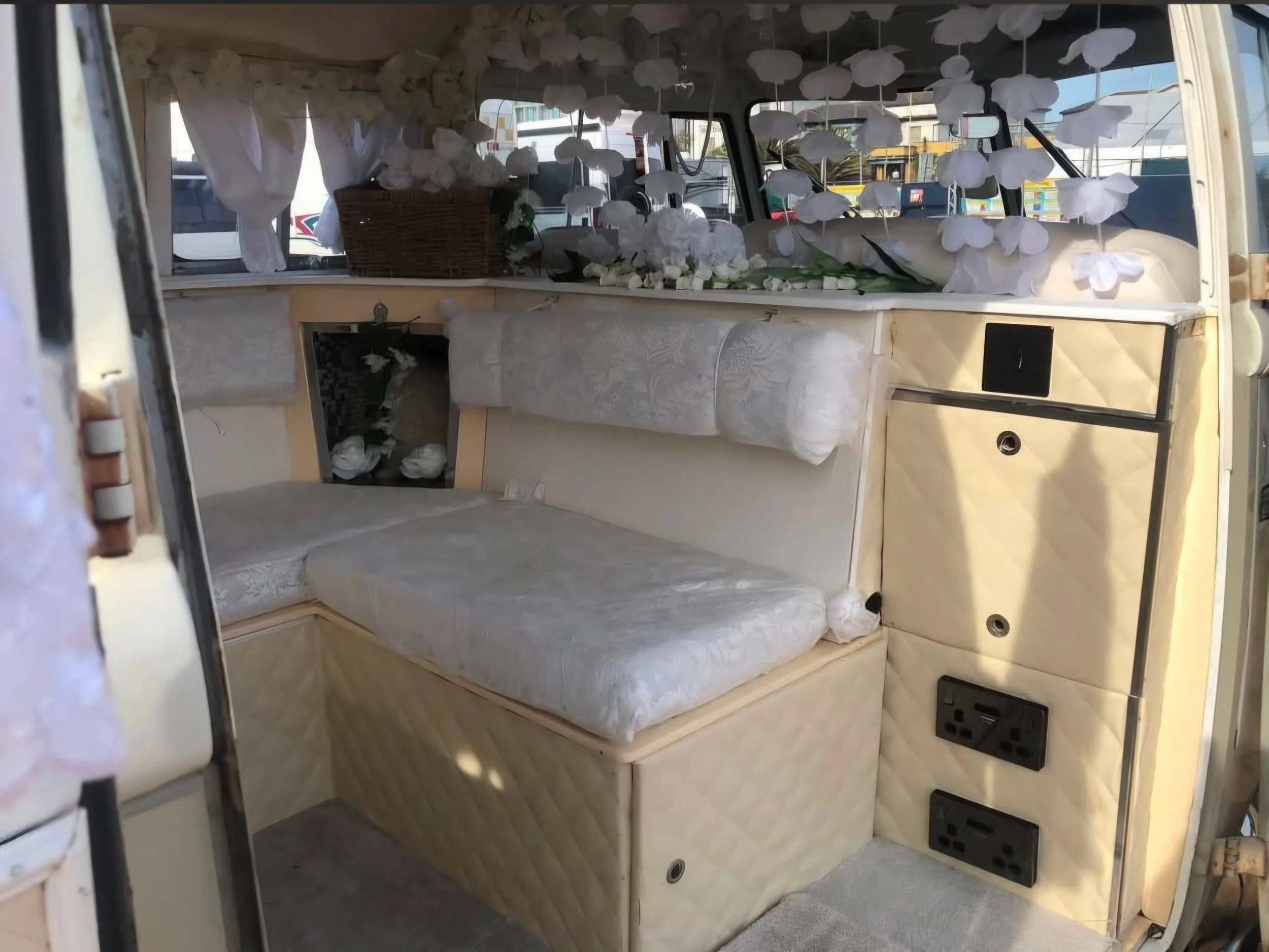 The interior of a vintage cream-colored van decorated with white paper flowers hanging from the ceiling and placed around the window. There is a wicker basket and a bouquet of white flowers on the dashboard. The van has a small kitchenette with a countertop and cabinetry, and various electrical outlets are visible at the bottom.
