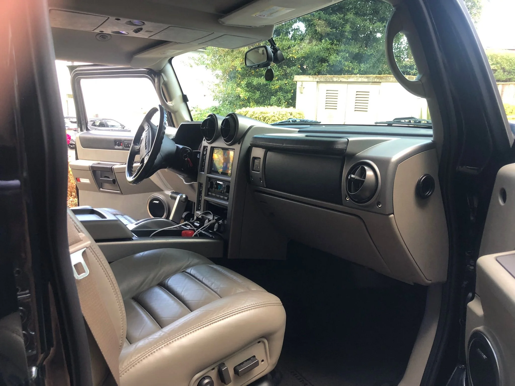 Inside view of a vehicle with beige leather seats, a dashboard with a touchscreen, and a view of trees and a building through the windshield.