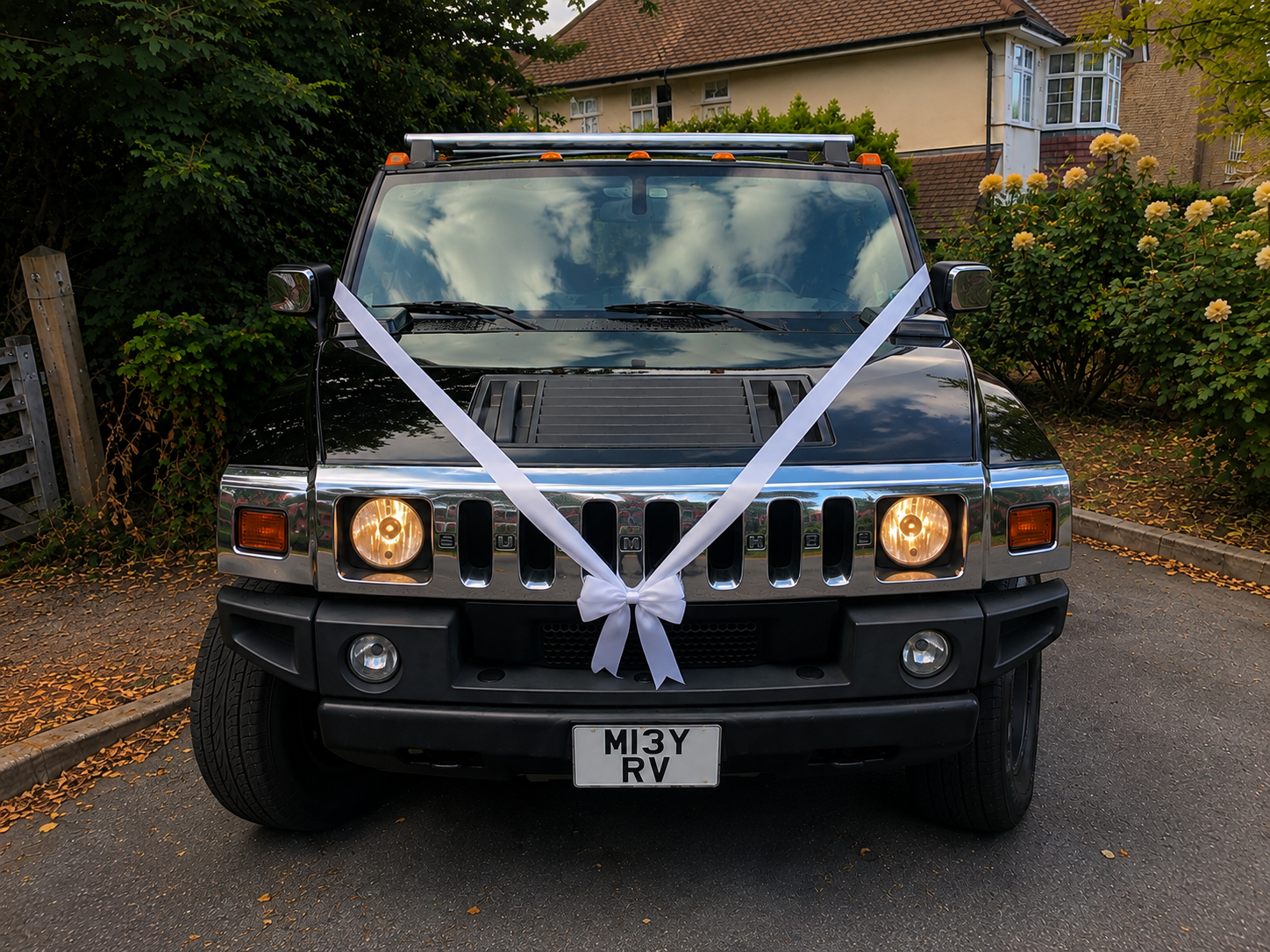 Front view of a black Hummer H2 vehicle decorated with a white ribbon tied in a bow on the front grille, parked on a driveway near bushes and a house in the background.