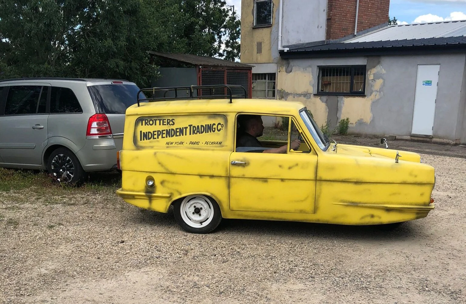 A small, yellow, homemade van with graffiti-style black spray paint on the side, parked on a gravel lot next to a silver SUV. The van has the text 'Trotters Independent Trading Co.' along with city names like New York, Paris, and Peckham. A person is sitting inside, holding a phone.