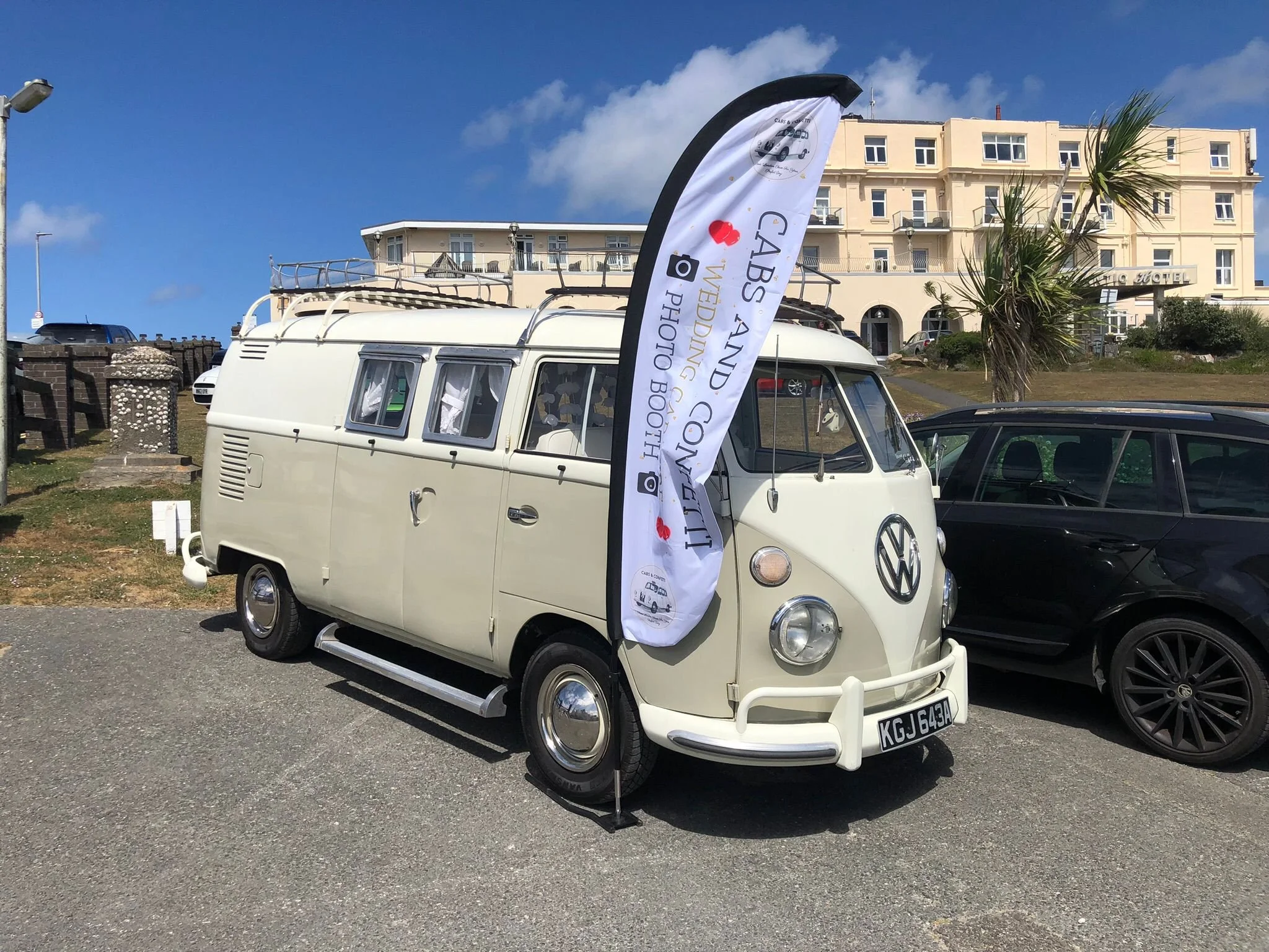 A vintage white Volkswagen van with a banner advertising a classic car and photo tour company, parked next to a black car on a paved lot. Buildings and a palm tree are visible in the background under a blue sky with some clouds.