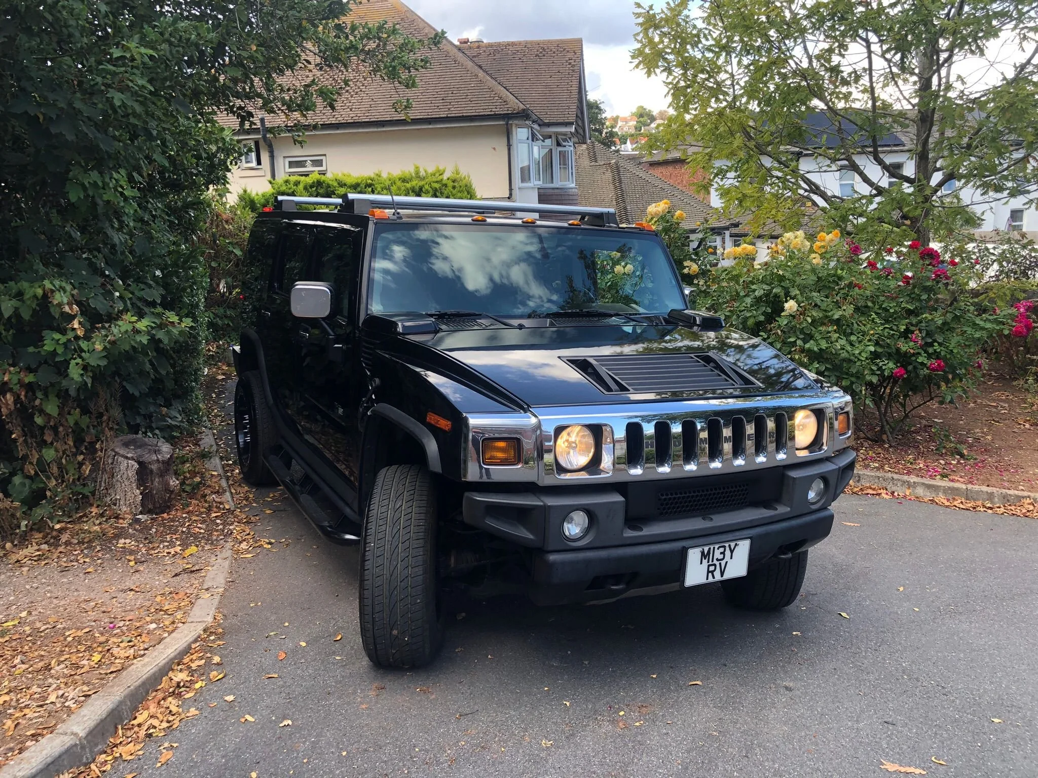 A black Hummer SUV parked on the side of a residential street next to a garden with a tree and flowering bushes, in front of houses with tiled roofs and a hillside in the background.