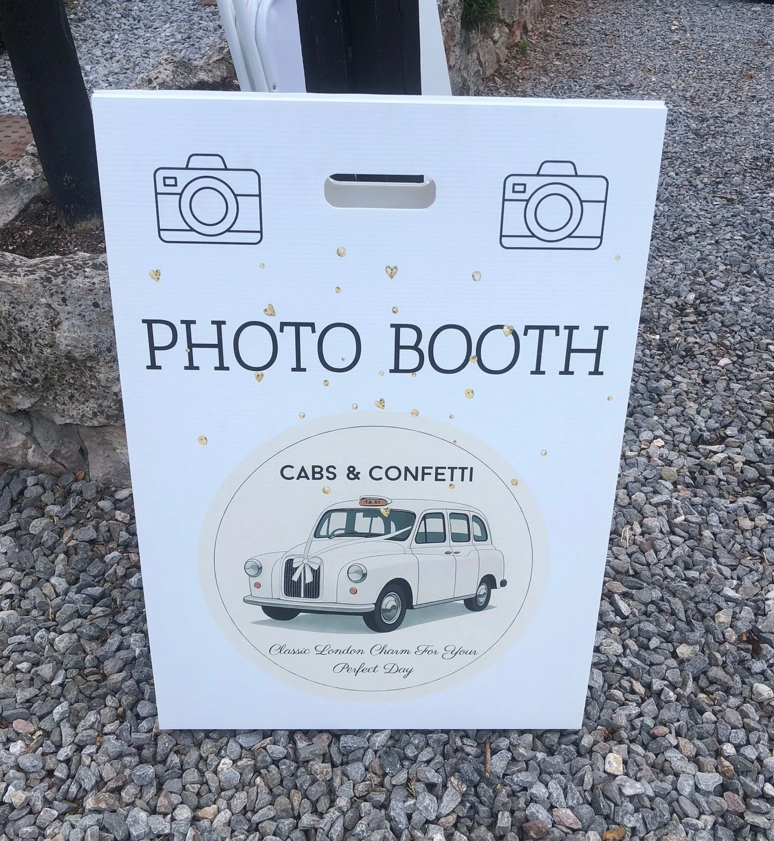 Sign for a photo booth featuring a vintage London taxi cab illustration, the words 'PHOTO BOOTH' and 'CABS & CONFETTI', and decorative gold hearts and dots, set on rocky ground.