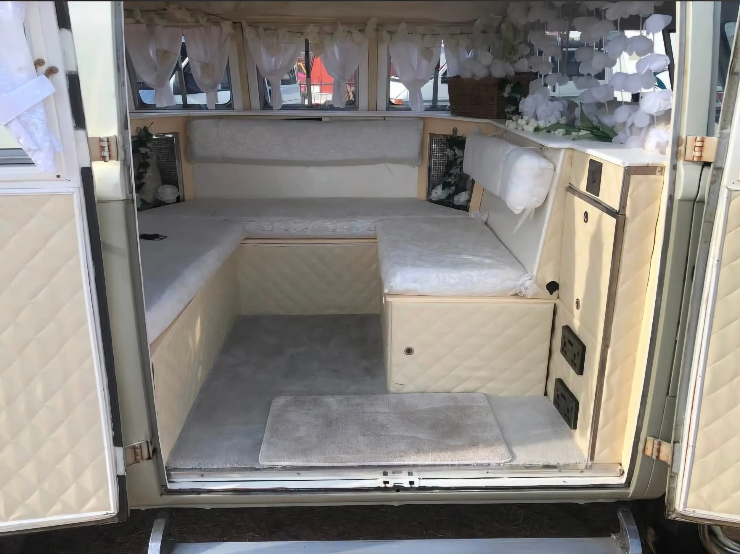 Inside a vintage camper with beige cushioned seating, small windows with white curtains, and a floral arrangement on the right side.
