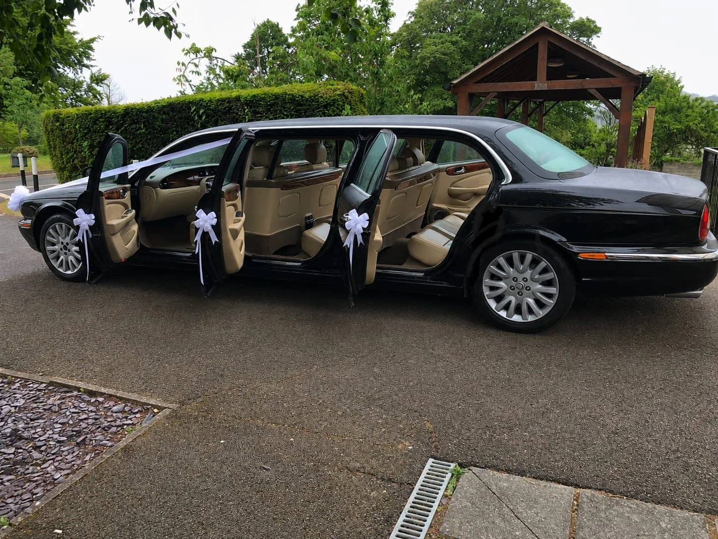 Black luxury sedan with four doors decorated with white bows, parked on a paved driveway surrounded by greenery and a wooden gazebo in the background.