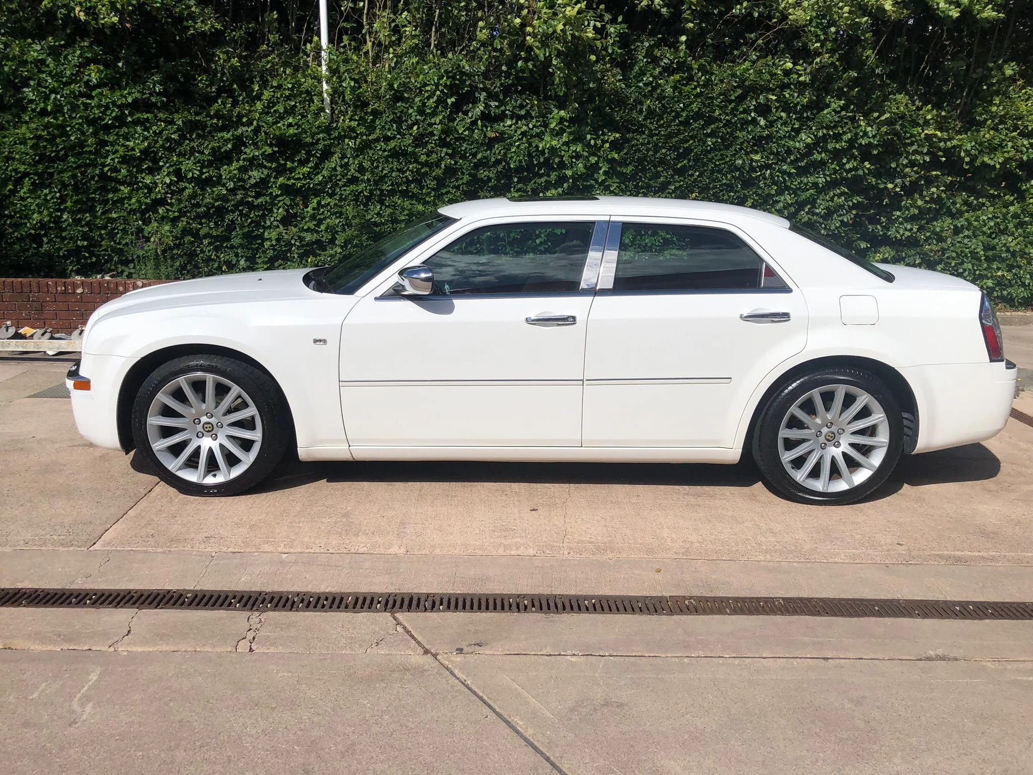 A white car parked on a street next to a brick sidewalk with a green hedge in the background.