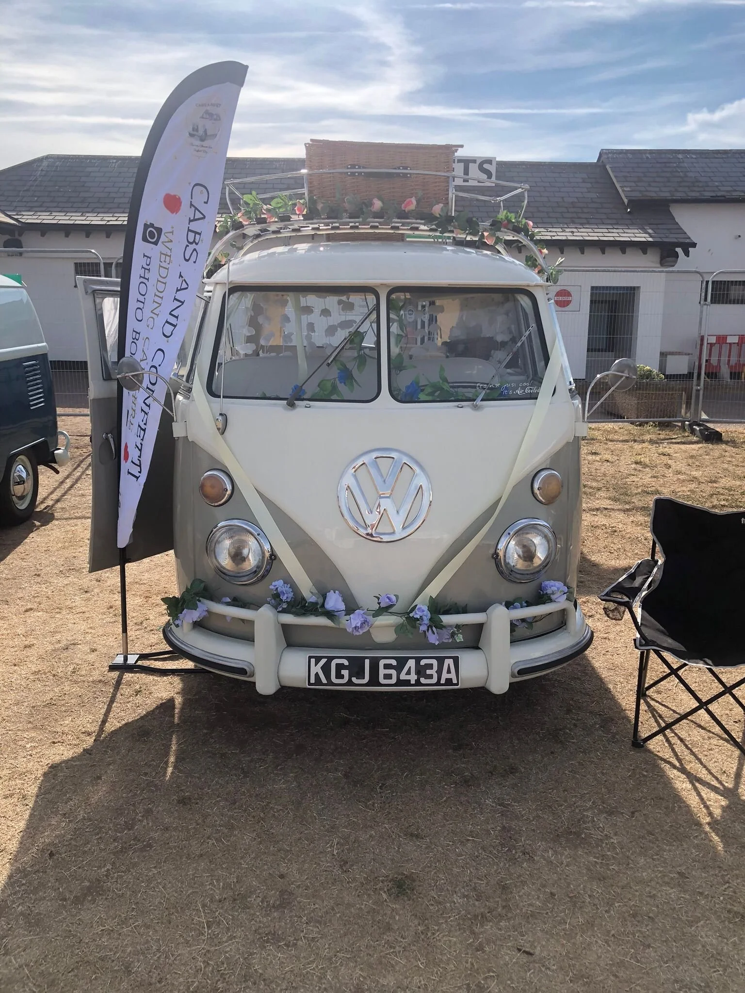 A vintage white Volkswagen van with floral decorations and a registration plate KGJ 643A, parked outdoors on a dirt ground with chairs nearby. A banner with the text 'CABS AND COUFT' is next to the van. There is a sign on the van's front saying 'It’s Air Cooler'. In the background, there is a building with a dark roof and some fencing.