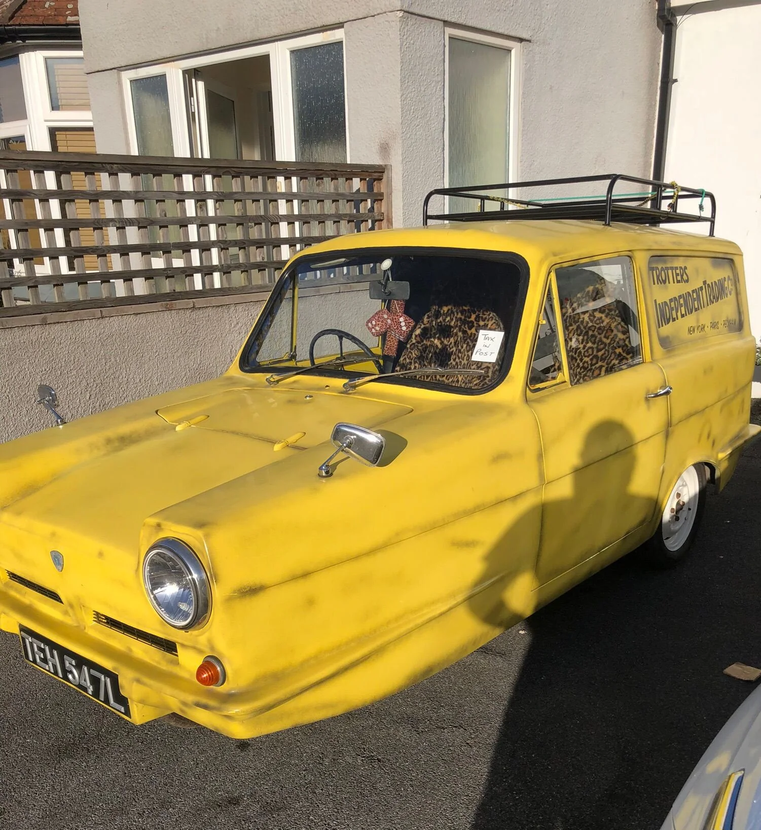 A vintage yellow car with a roof rack parked outdoors in front of a residential building, with a wooden fence and a large window visible in the background.