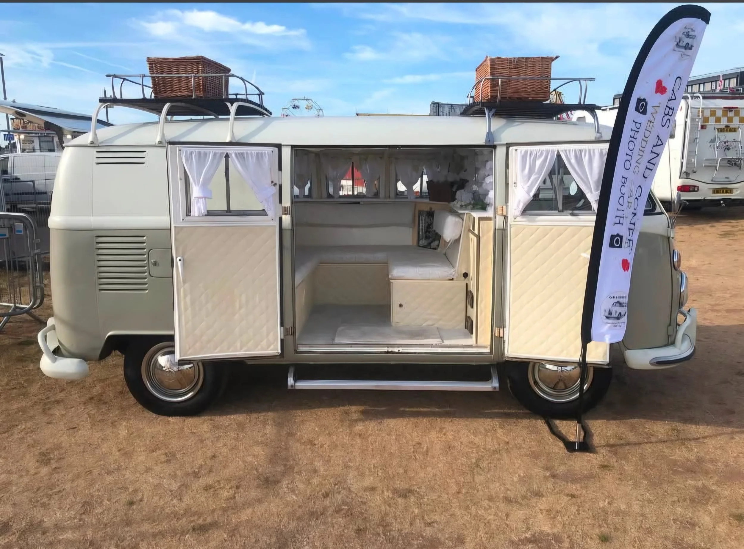A vintage white camper van with a diamond-patterned exterior, open side door revealing a cozy interior, and white curtains on the windows. The van has roof racks with wicker baskets on top. A black and white flag stands next to the van advertising a photography booth at an outdoor event.