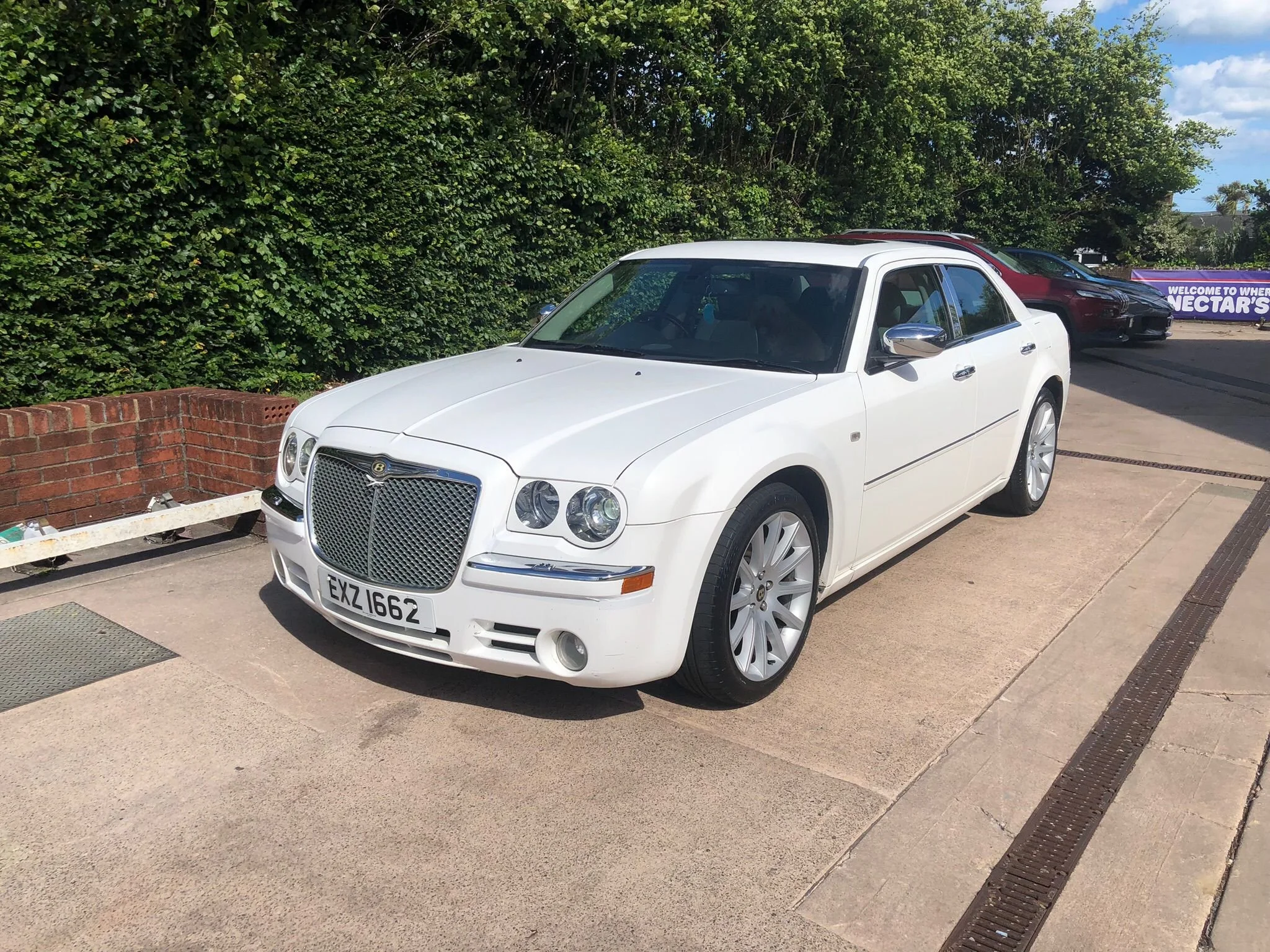 White luxury car parked on a driveway next to a red brick wall and green bushes. Additional cars are parked nearby, and a purple sign in the background reads "Welcome to where Nectar's."