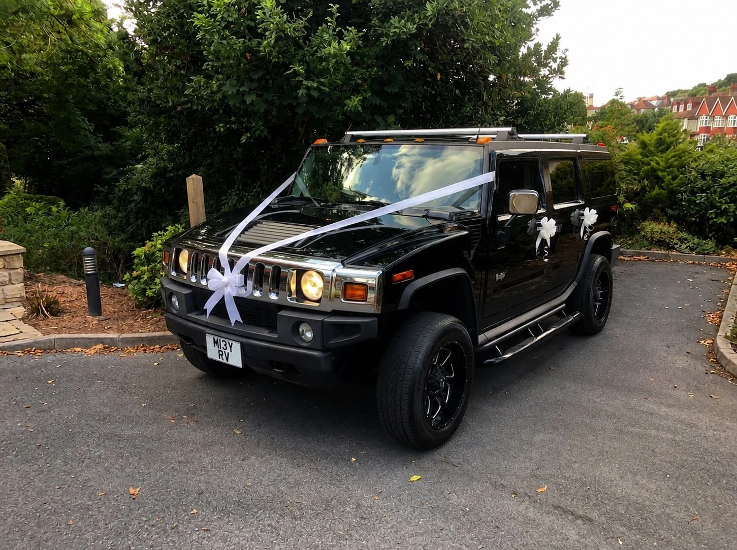 A black Hummer H3 decorated with white ribbons and bows, parked on a paved driveway near greenery, likely for a wedding or special event.