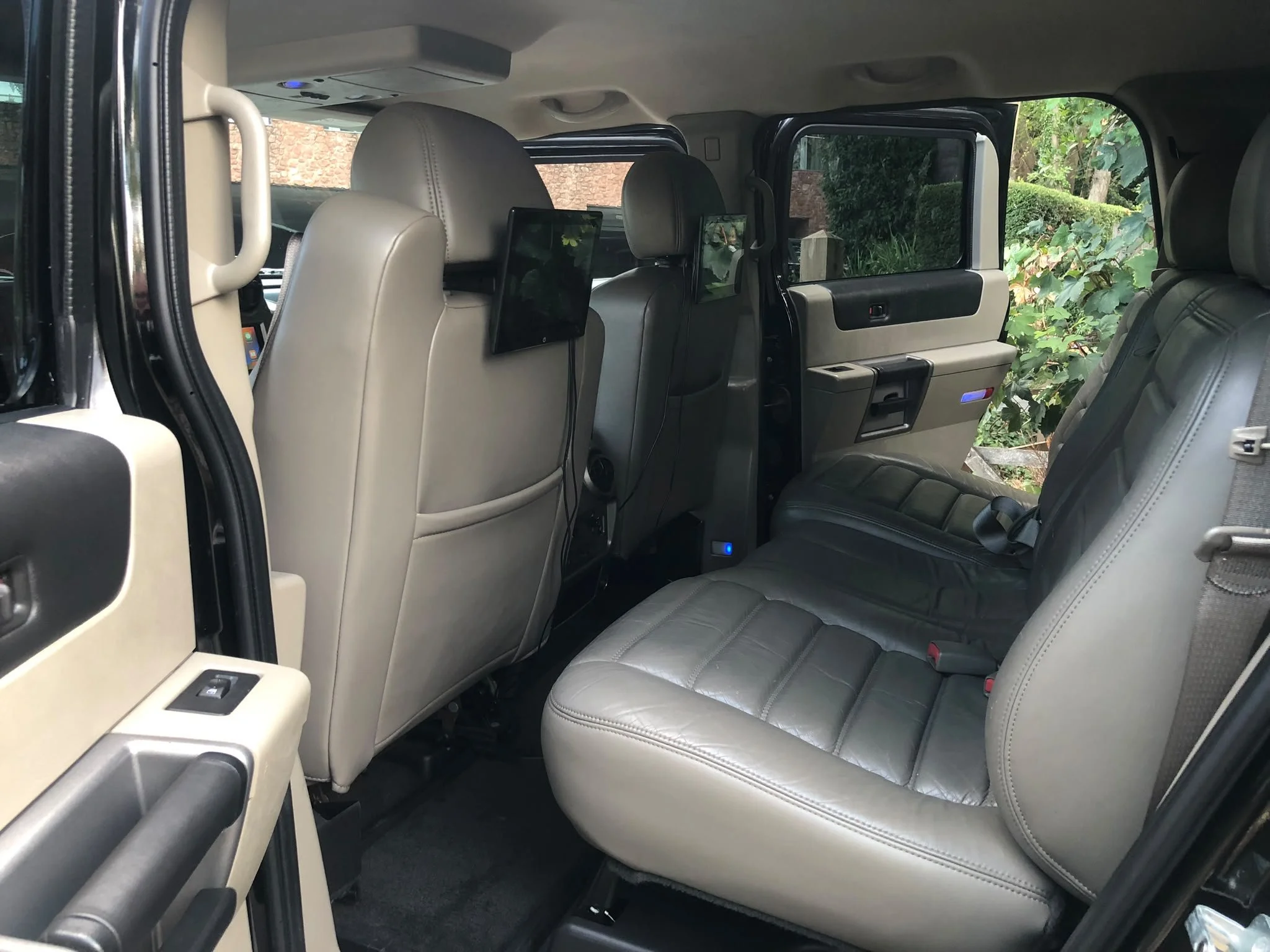 Interior of a vehicle showing beige leather seats, headrest-mounted screens, and a side window with a view of greenery and bushes outside.