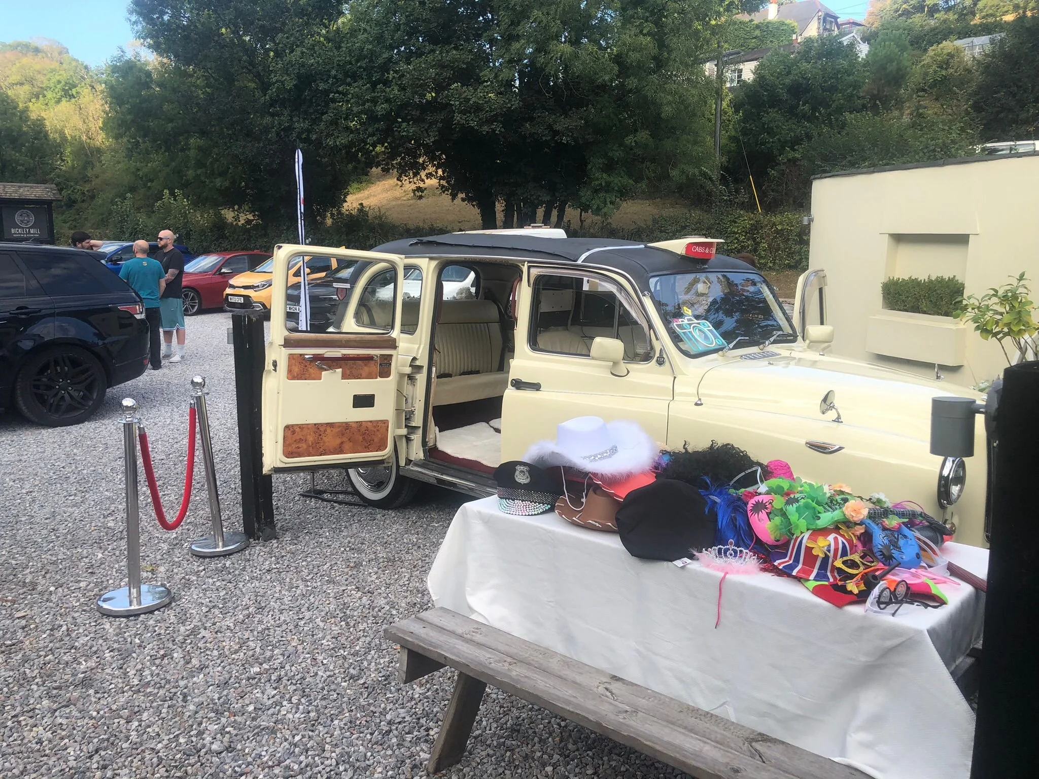 Outdoor flea market or yard sale with vintage car, tables with hats, sunglasses, accessories, and costumes, surrounded by parked cars and trees.