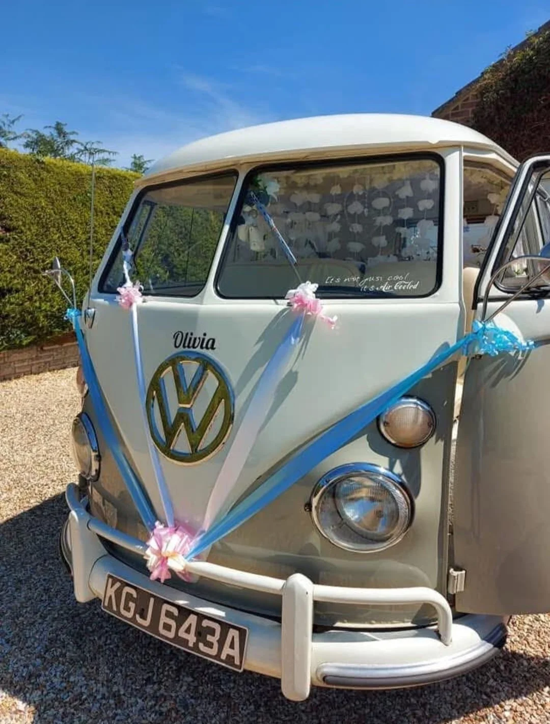Vintage Volkswagen van decorated with pink, blue, and white ribbons and bows, with the name 'Olivia' and a gold VW logo on the front, parked on gravel under a clear blue sky.