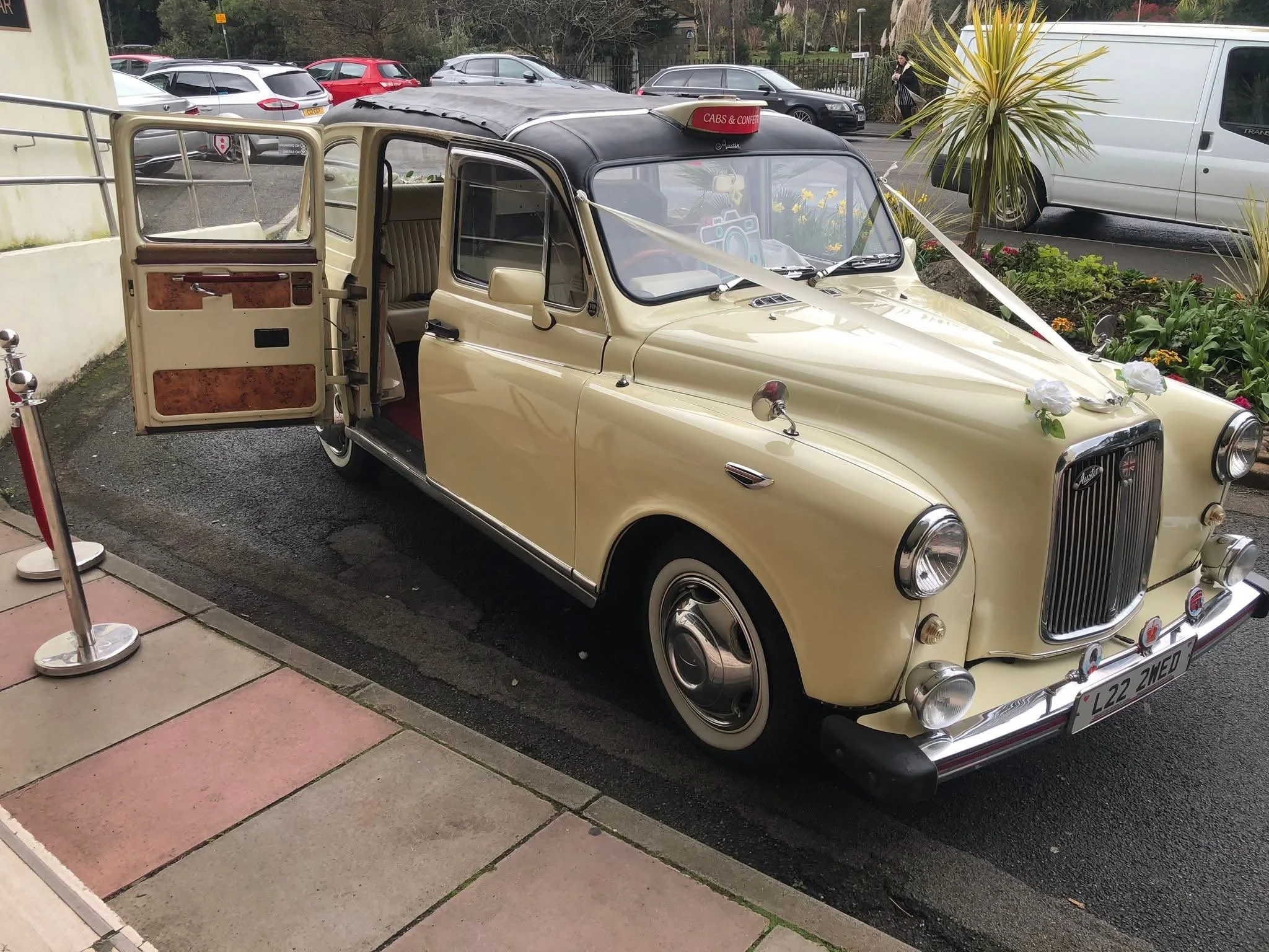 A vintage yellow and black car decorated with ribbons and flowers, parked outside a building with other cars and people in the background.