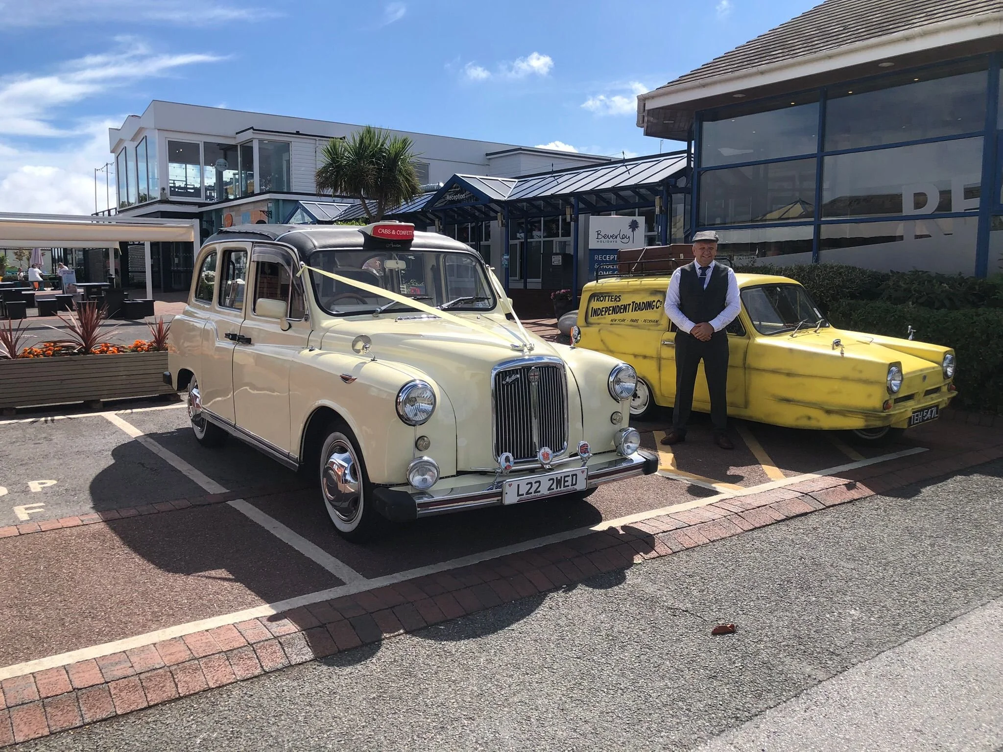 Two vintage taxis parked in front of a resort with a man standing next to a yellow one. The resort building has a glass entrance with signs for reception and holiday rentals, blue frames, and a palm tree nearby. The sky is partly cloudy with a few clouds.