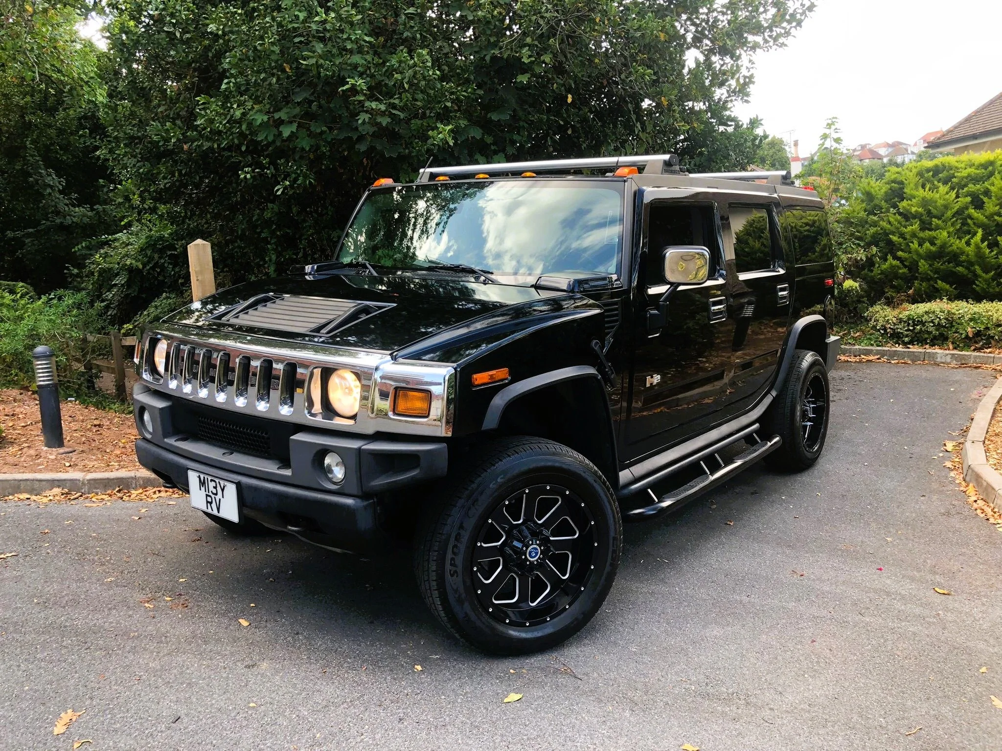 A black Hummer vehicle parked in a parking lot surrounded by green trees and shrubs, with residential houses in the background.