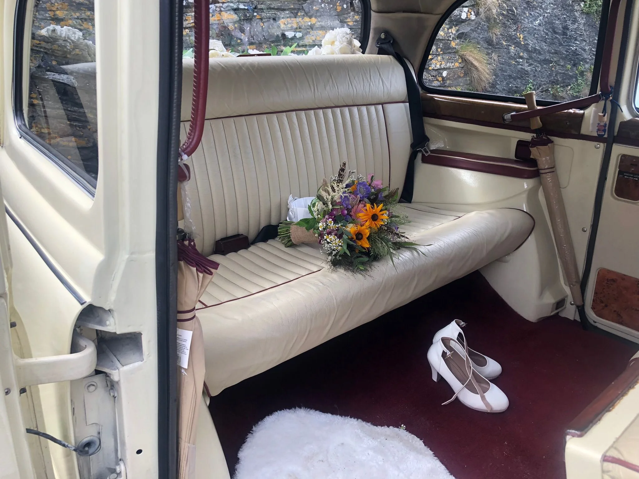 Inside a vintage white car with a bench seat, a bouquet of flowers resting on the seat, a pair of white shoes on the floor, and a window showing rocky terrain outside.