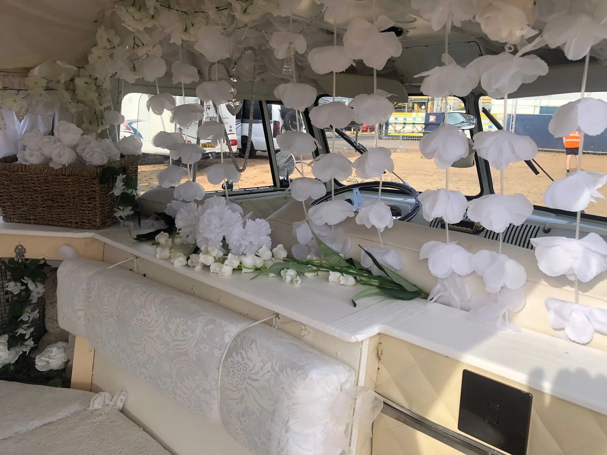 A decorated interior space with white floral decorations hanging from the ceiling, and a wicker basket filled with white flowers on a table covered with a lace tablecloth. The scene appears to be set up for an event, with a parking lot visible through the windows in the background.