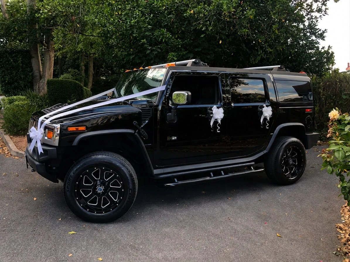 A black SUV decorated with white ribbons and bows, likely for a wedding, parked on a paved driveway with greenery and flowers in the background.