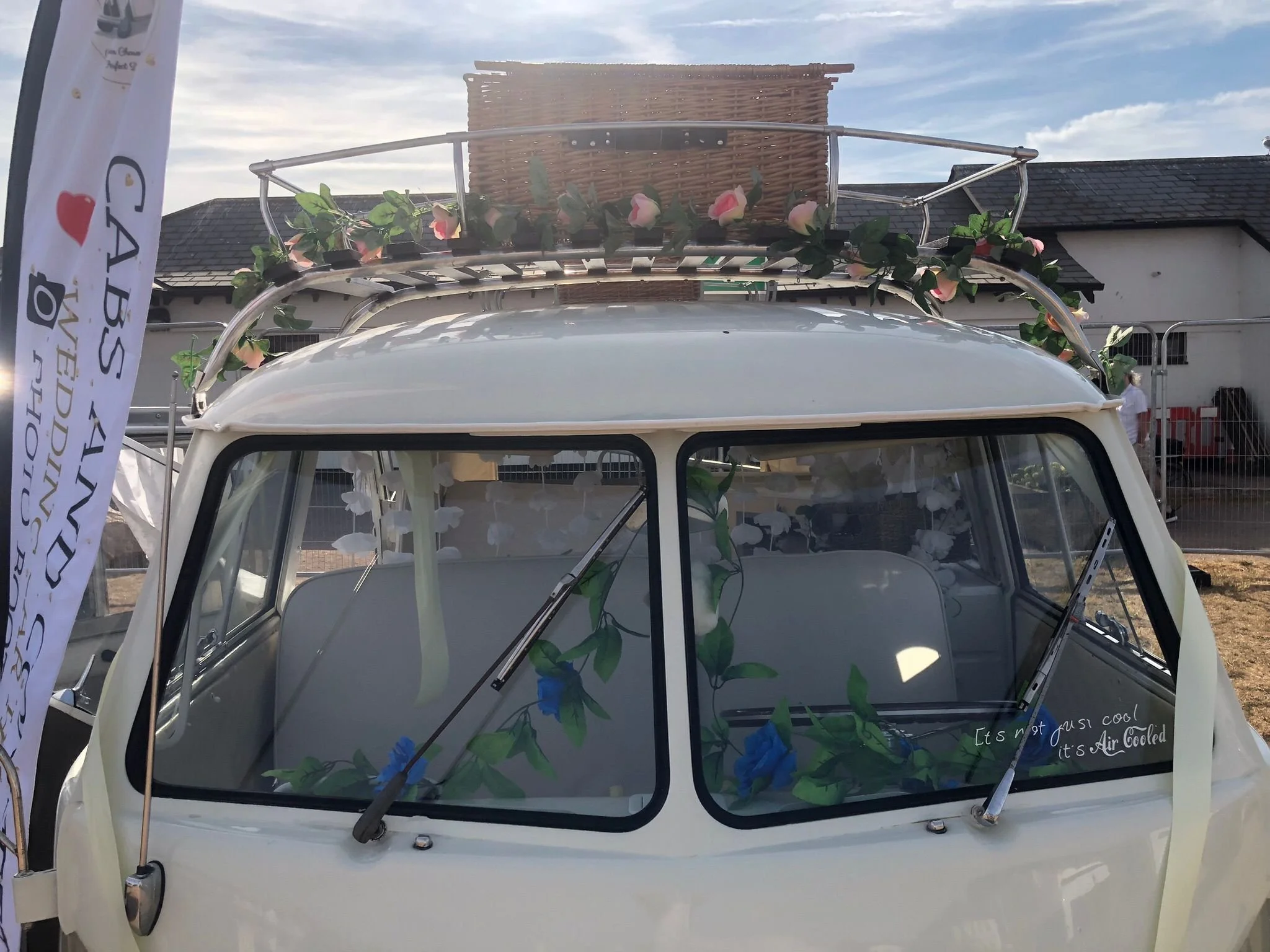 A close-up of a vintage white vehicle with a roof rack decorated with pink flowers and green leaves. The vehicle has slogans written on the windshield, including 'It's not just a cool car,' and a flag or banner with logos and text on the left side.