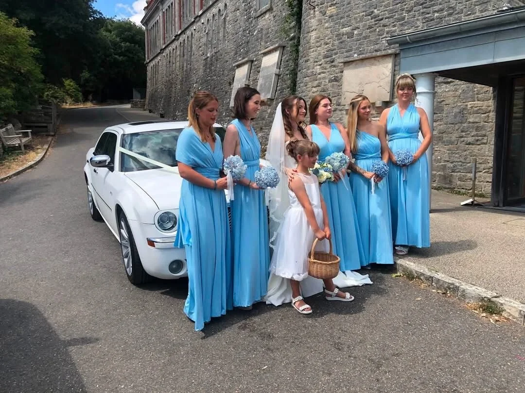 A bride and six bridesmaids in blue dresses, standing next to a white car decorated with ribbon, outside a stone building, with a young girl holding a basket in front of them.