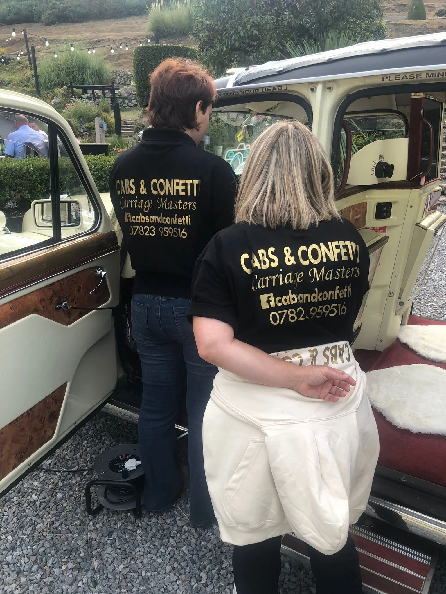 Two women wearing black jackets with yellow text reading 'Cabs & Confetti Carriage Masters' and contact details, standing next to a vintage-style carriage with a cream and brown interior and exterior, on a gravel surface in an outdoor setting with greenery and string lights in the background.