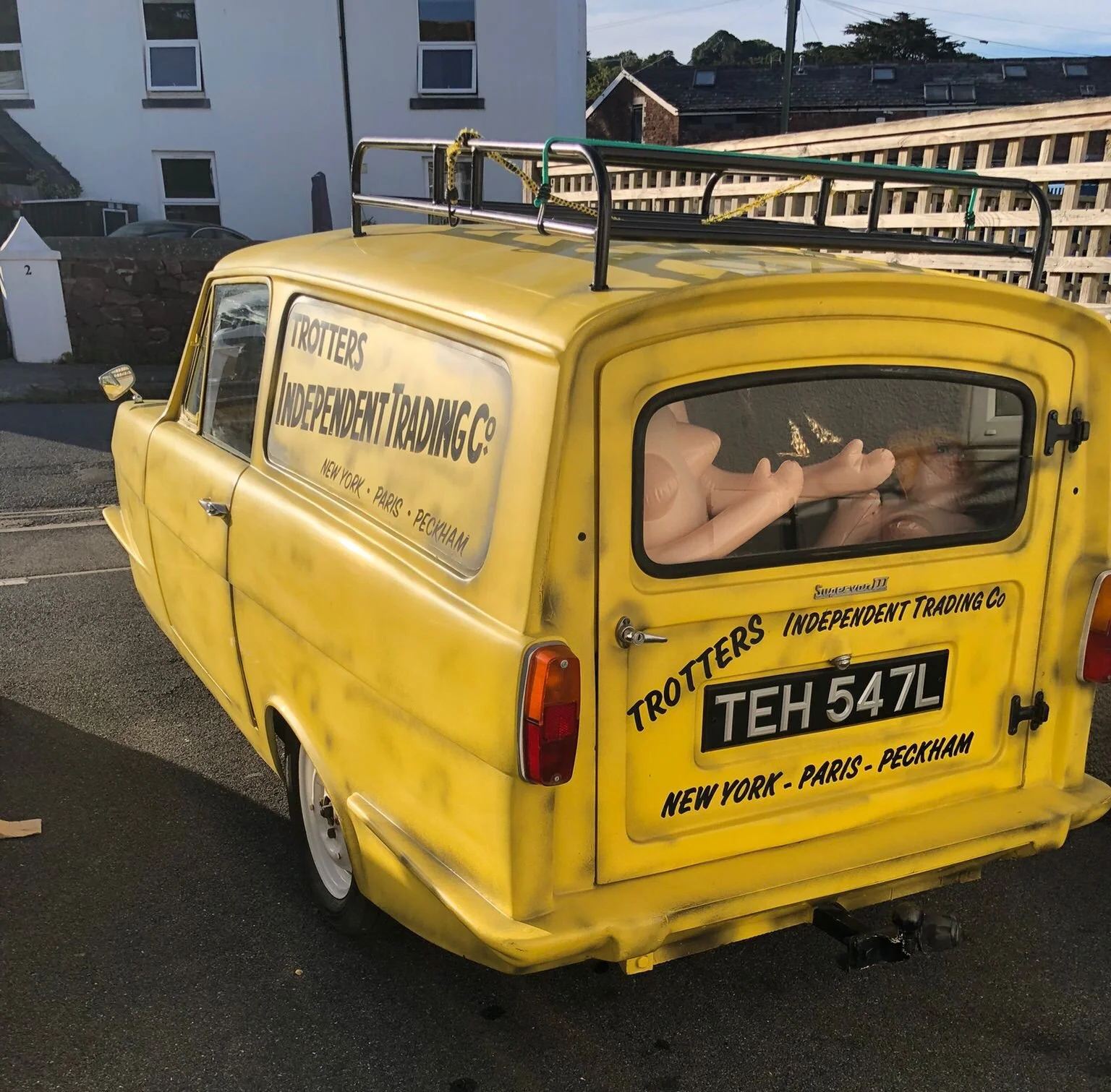 Yellow vintage van with signs reading "Trotters Independent Trading Co" and list of cities, parked on street in front of houses, with luggage rack on top, rear window displaying action figures inside.
