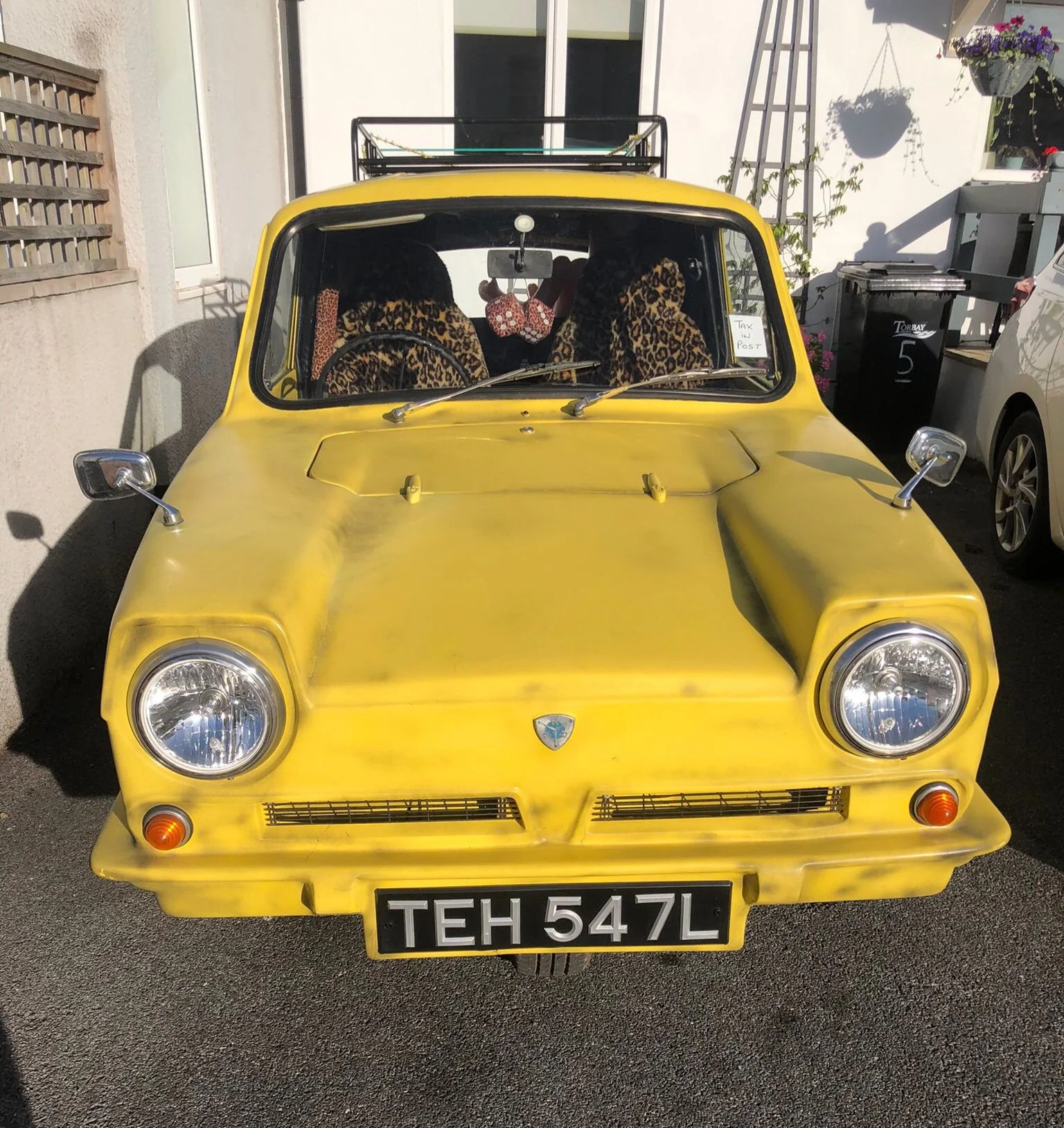 A yellow vintage three-wheeled car with leopard print seat covers, a roof rack, and a police-style license plate reading 'TEH 547L', parked on asphalt in front of a building with a small window and a shed.