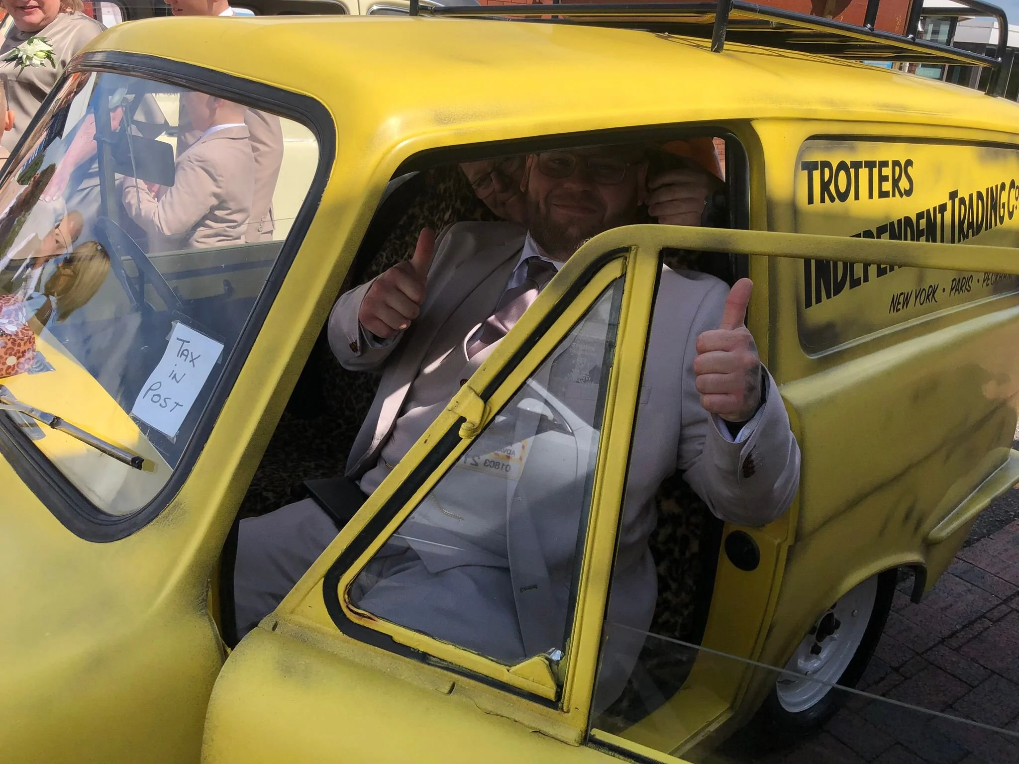 A man smiling and giving a thumbs up from the driver side of a bright yellow three-wheeled vehicle, with other people and a building in the background.