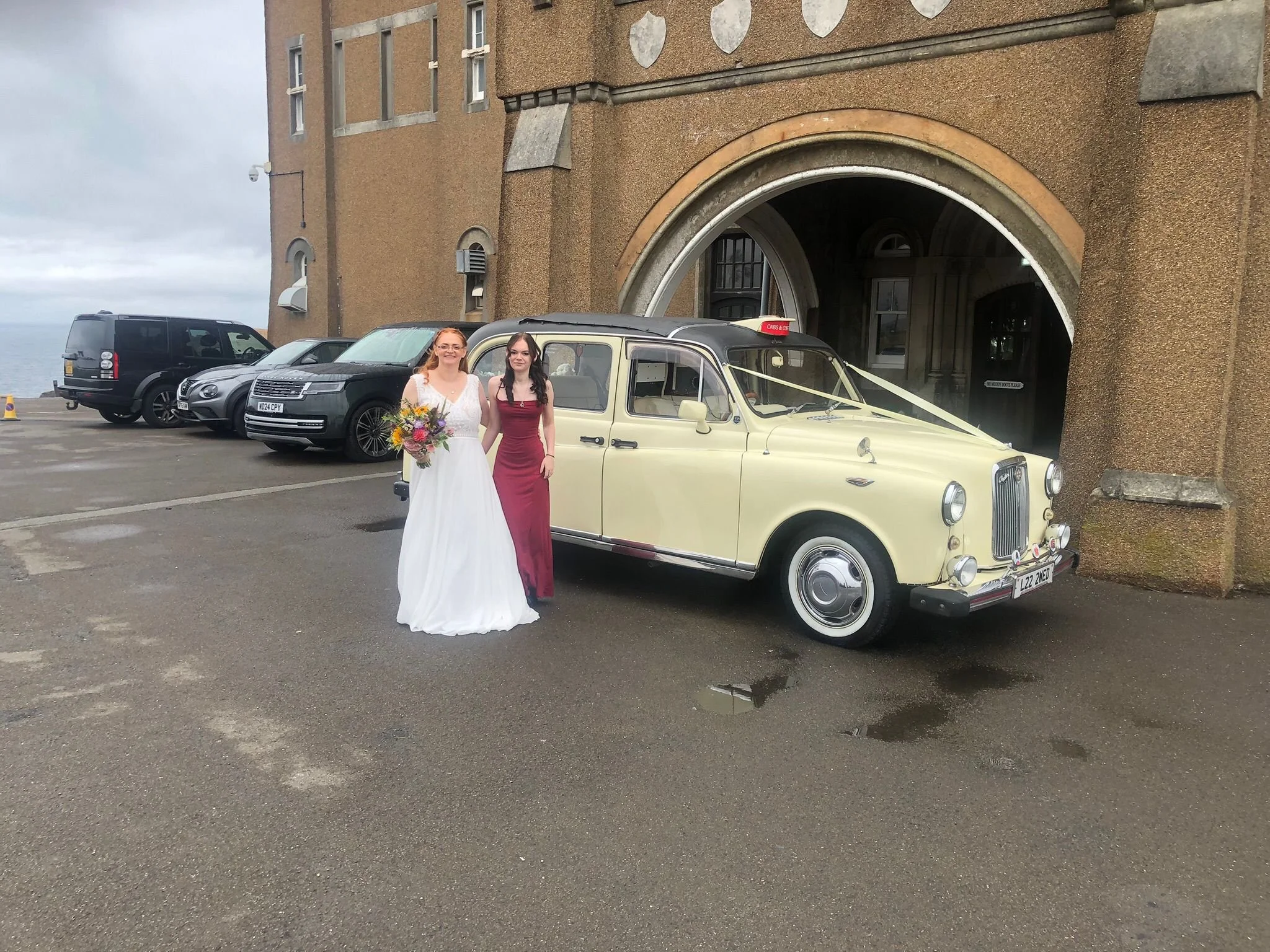 Two women in wedding attire standing next to a vintage taxi cab in front of a building with an archway entrance. One woman wears a white wedding dress holding a bouquet, and the other wears a red dress.