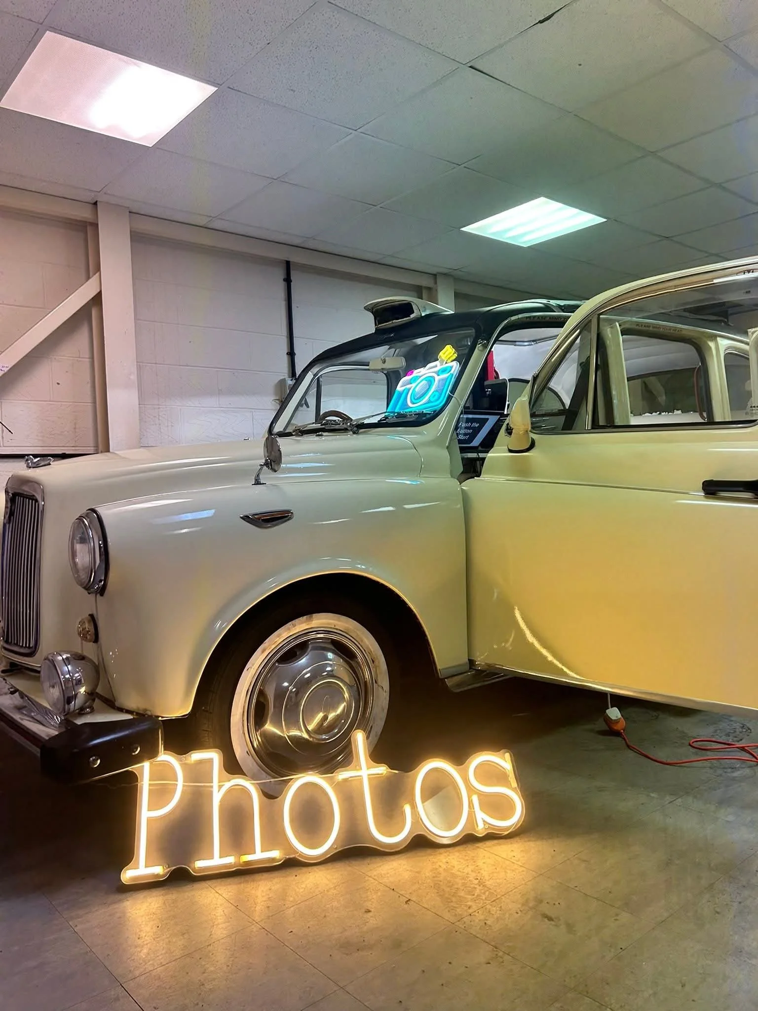 A vintage white car parked indoors with a neon sign that reads 'Photos' in front of it, and neon lights on the ceiling.