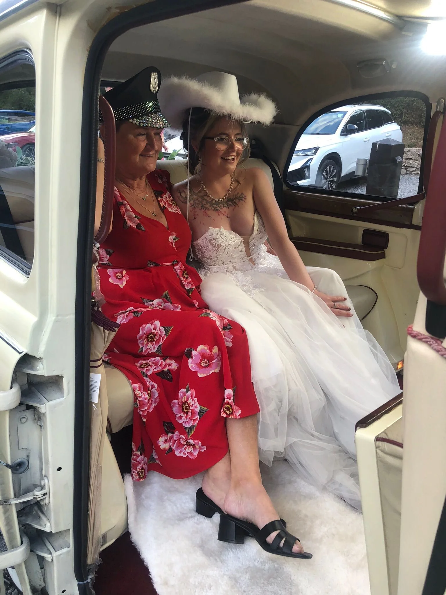 A woman in a white wedding dress and a woman in a red floral dress sitting inside a vintage car, both wearing fancy hats, with the woman in the wedding dress smiling.