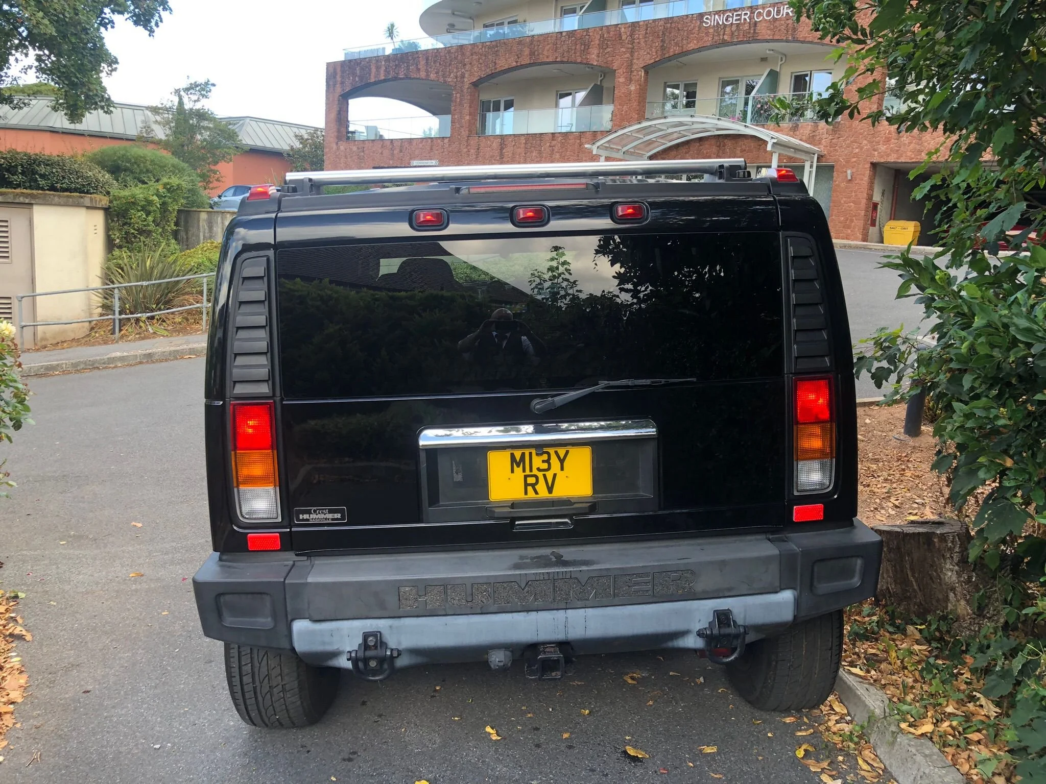 Rear view of a black Hummer SUV with a yellow license plate reading 'M13Y RV,' parked on the side of a street near a building with a brick and white exterior and balconies. Surrounding greenery and fallen leaves are visible, and a reflection of the photographer is seen on the vehicle's rear window.