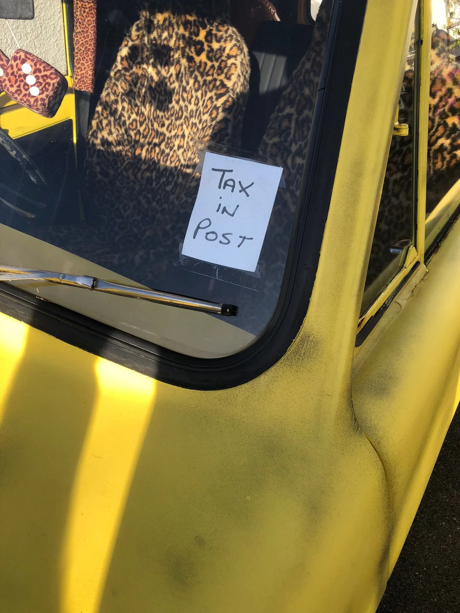 A yellow vehicle with a leopard print interior visible through the windshield. There is a handwritten sign taped to the inside of the windshield that reads 'TAX IN POST.' The vehicle's side mirror is also leopard print.