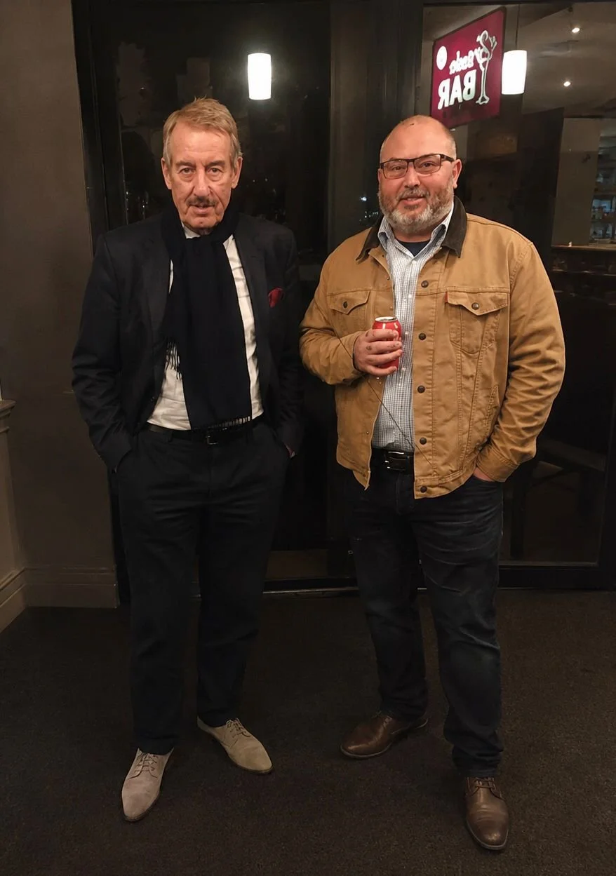 Two men standing indoors. The man on the left wears a black suit, white shirt, black scarf, and beige shoes. The man on the right wears glasses, a tan jacket, checked shirt, dark jeans, brown shoes, and holds a red can. There is a sign that reads 'Bar' in the background.