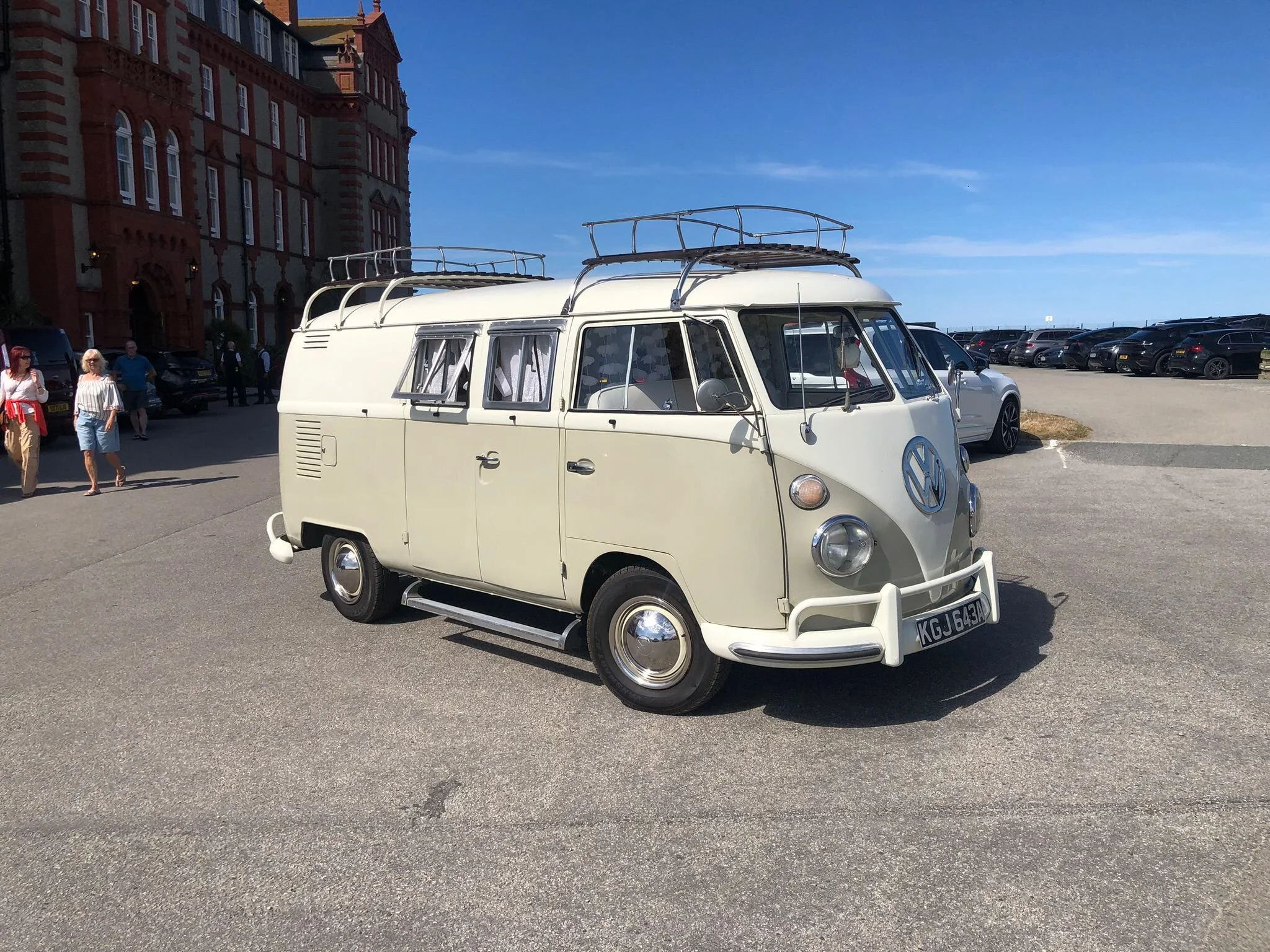 A vintage cream-colored Volkswagen camper van with a roof rack parked in an outdoor parking lot on a sunny day. There are several people walking around and a large brick building in the background.