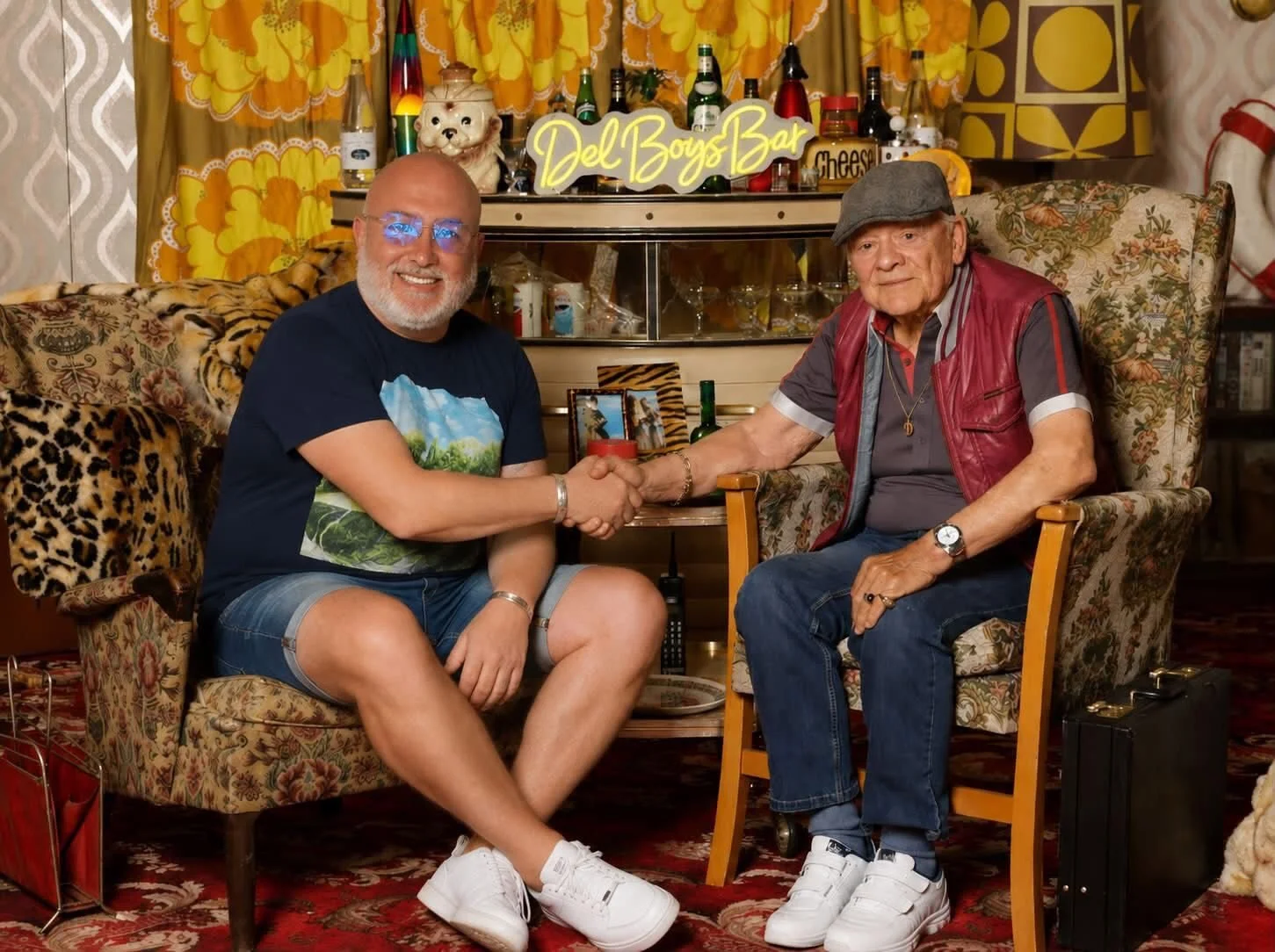Two men sitting in vintage-style living room, shaking hands. One appears to be a young man with glasses, casual clothing, and bald; the other is an older man with a gray cap and red vest. Behind them is a bar with a yellow neon sign that says 'Del Boys Bar,' and colorful curtains.