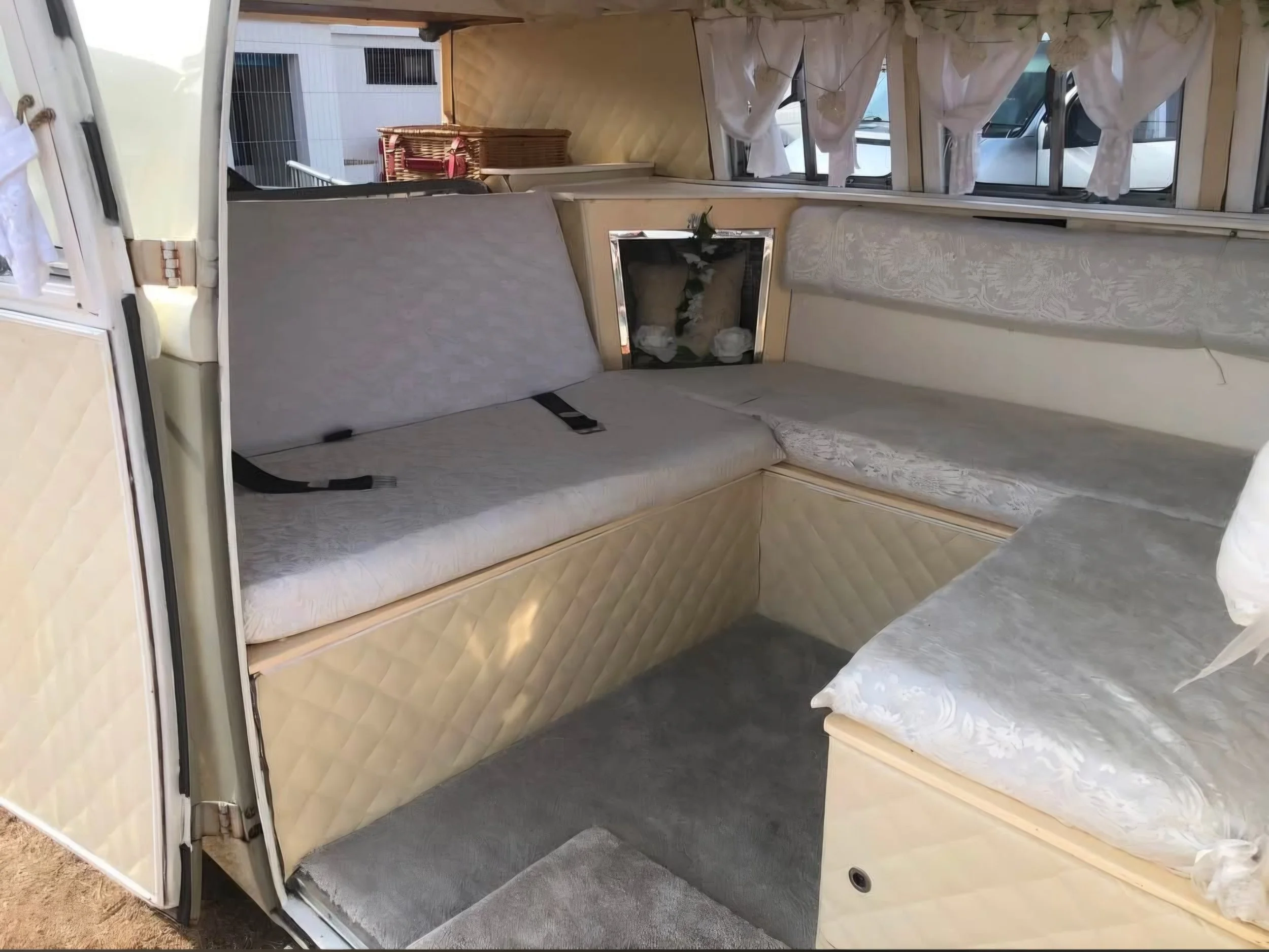 Interior of a vintage camper with cream-colored cushioned seating along the walls and a small fireplace with a mirror above it. There are windows with white curtains and various woven baskets and items on a table.